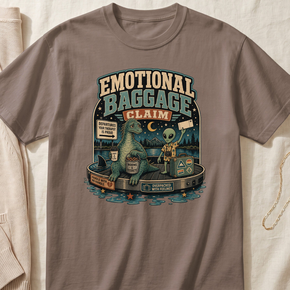 Graphic tee with "Emotional Baggage Claim" text, dinosaur, alien, and luggage.