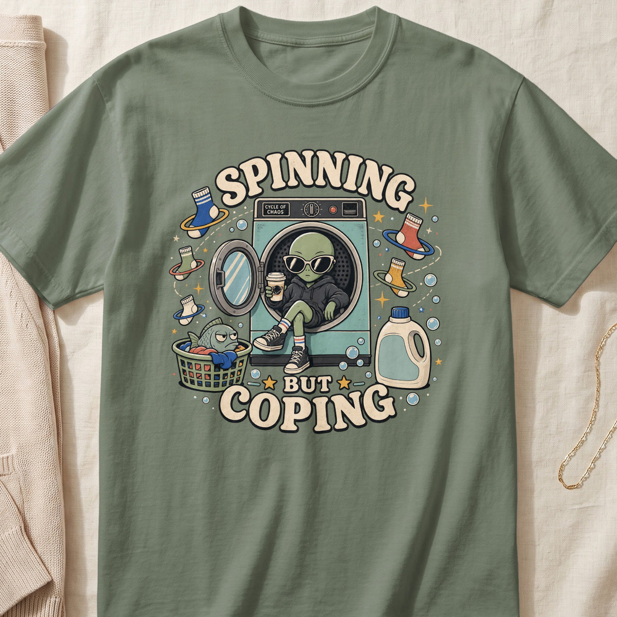Green t-shirt graphic of alien sitting in washing machine with "Spinning but coping" text.