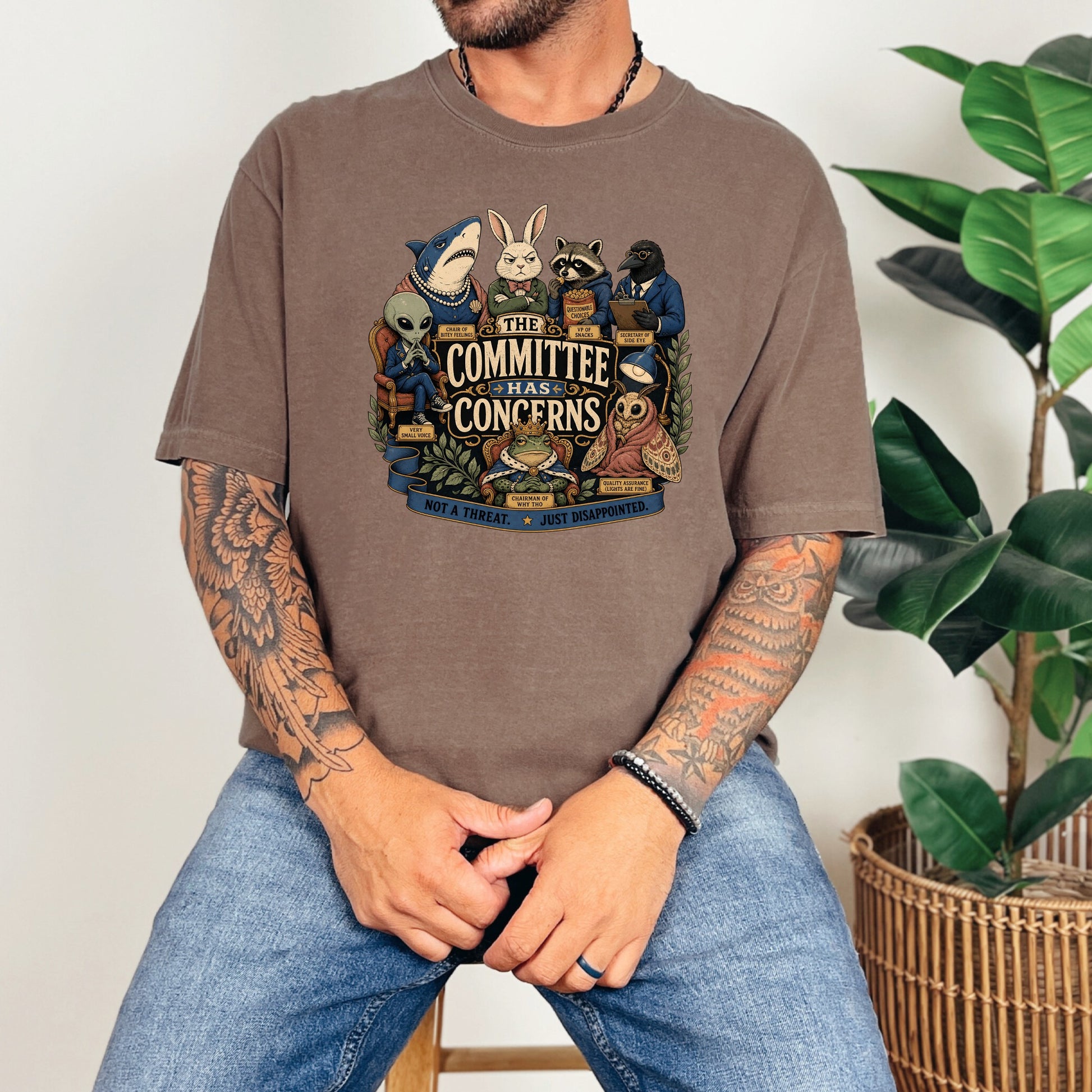 A tattooed man wearing a brown t-shirt featuring a cartoon committee graphic with anthropomorphic animals.