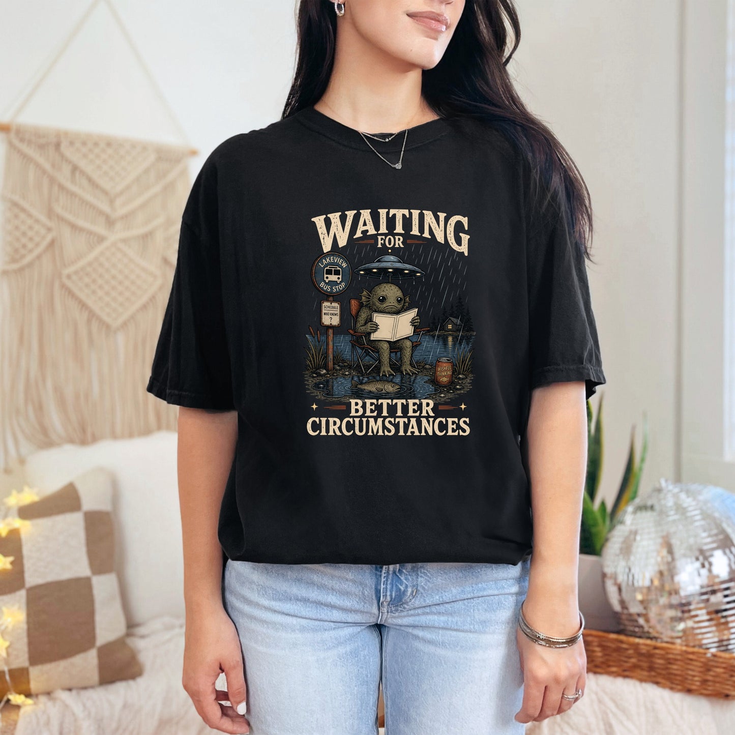 T-shirt graphic: creature under UFO at bus stop reading, "Waiting for better circumstances."