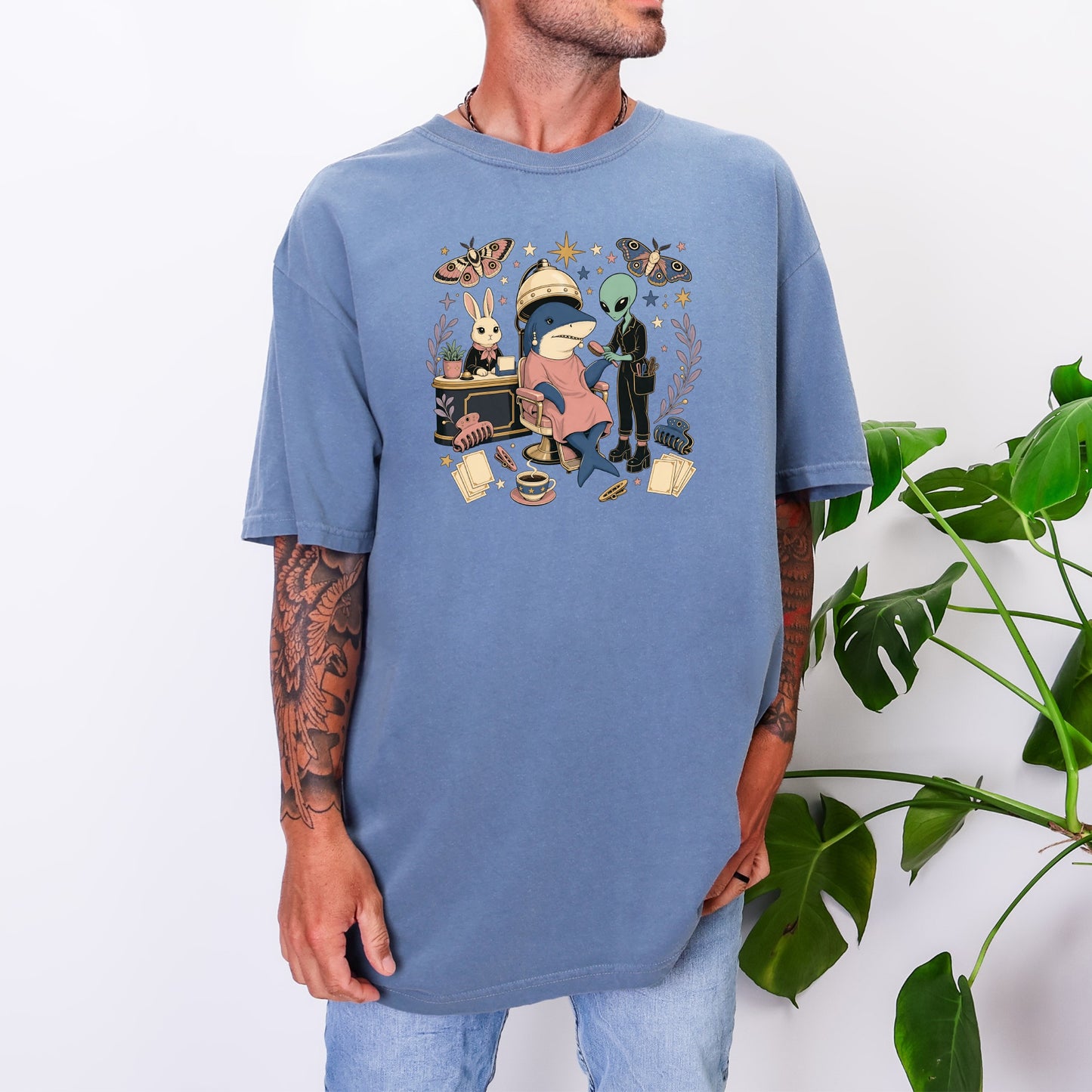 A person wearing a blue oversized t-shirt printed with a whimsical salon illustration featuring a shark, alien, and rabbit.
