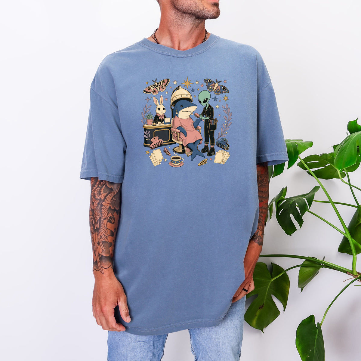 A person wearing a blue oversized t-shirt printed with a whimsical salon illustration featuring a shark, alien, and rabbit.