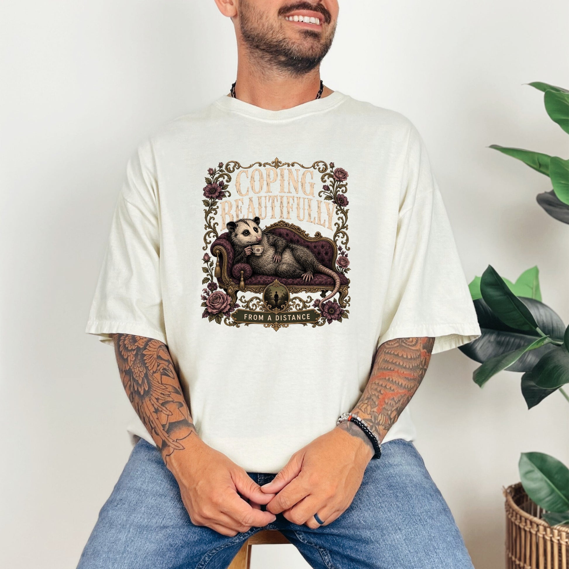 Cream t-shirt with a graphic of an opossum lounging and text "Coping Beautifully From A Distance."