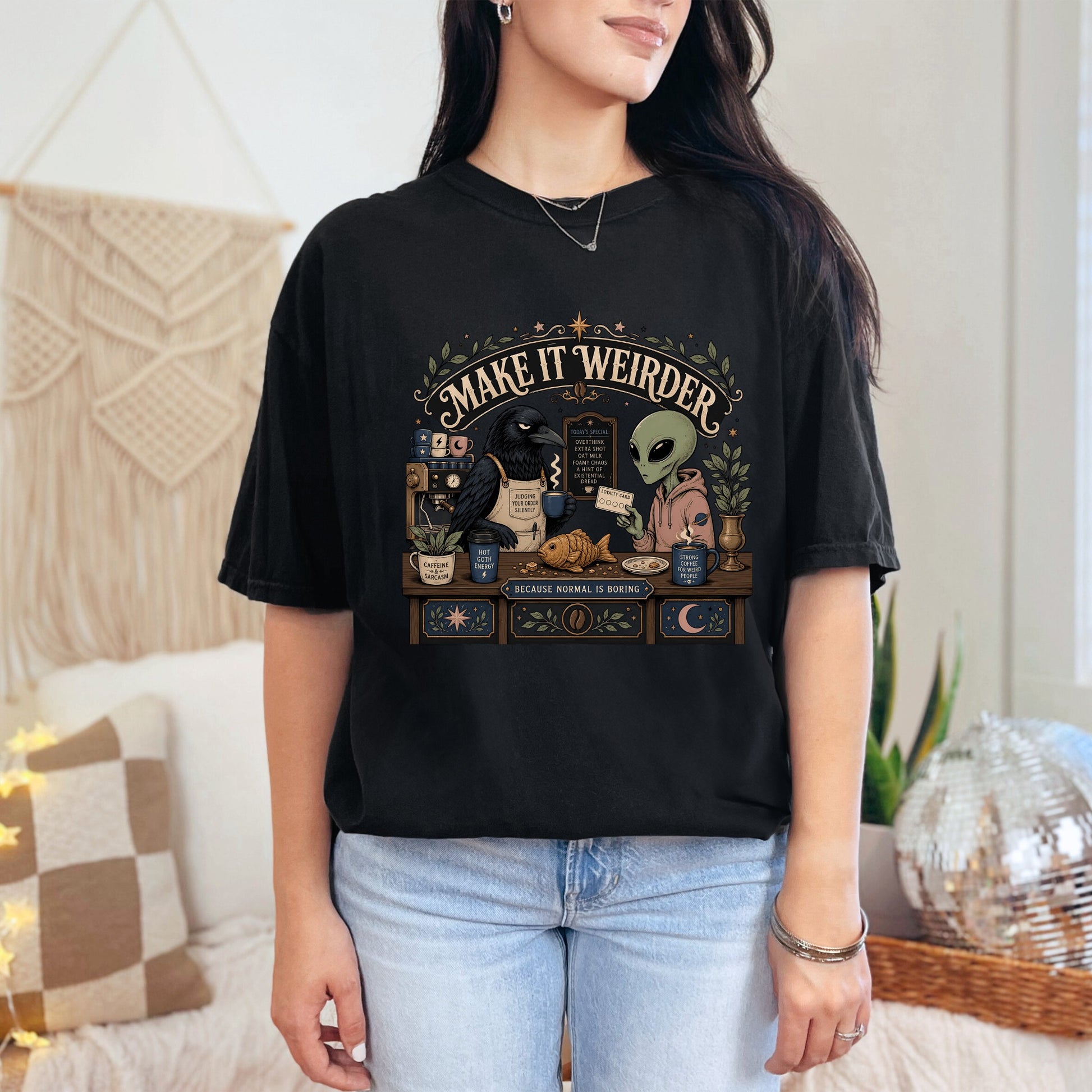 Black t-shirt with alien and crow at coffee counter, "Make It Weirder" text.