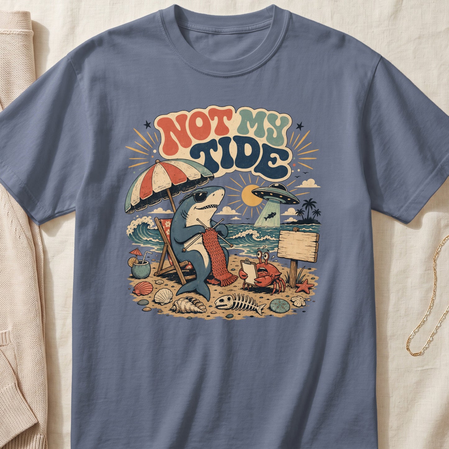 Cartoon shark knitting on beach with crab, UFO, and text "NOT MY TIDE".