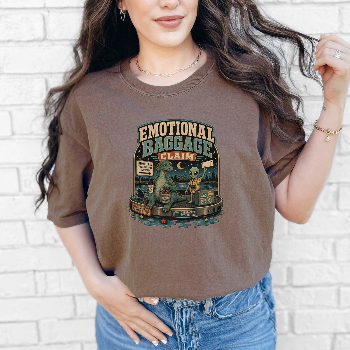 Brown t-shirt with "Emotional Baggage Claim" graphic of dinosaur, alien, and suitcase.