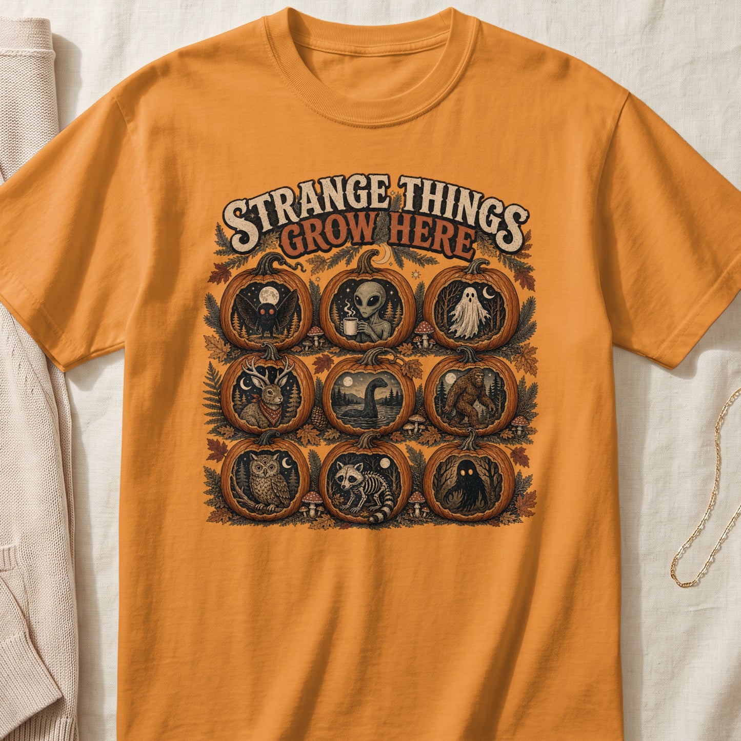 Orange t-shirt with "Strange Things Grow Here" and nine pumpkin designs featuring cryptids.