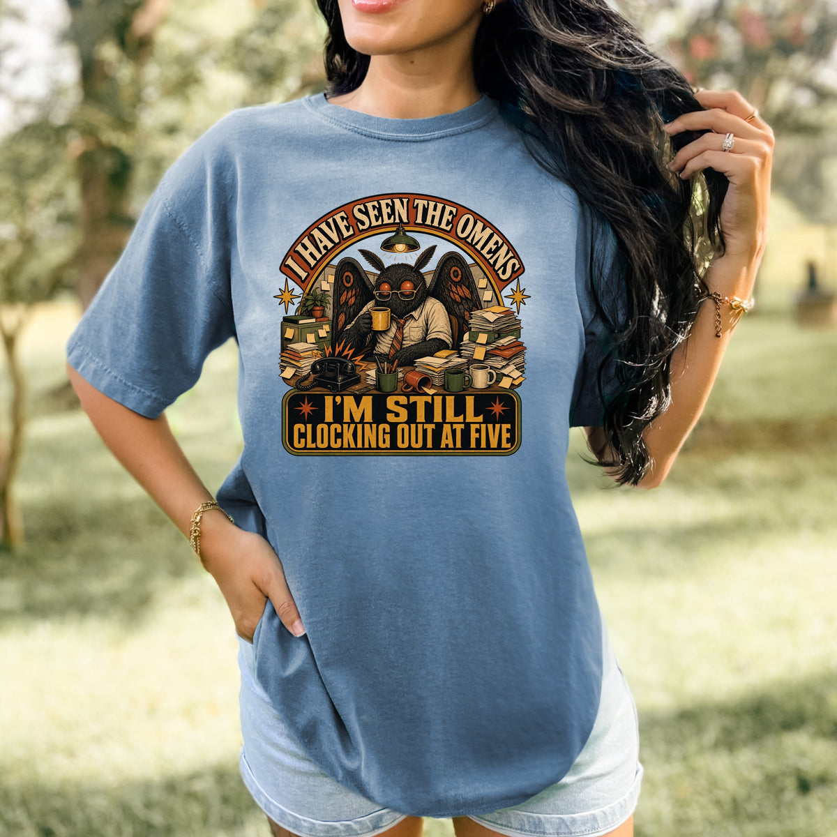 Graphic on a blue t-shirt shows a mothman character with wings, glasses, and a tie, text says "I HAVE SEEN THE OMENS I'M STILL CLOCKING OUT AT FIVE"
