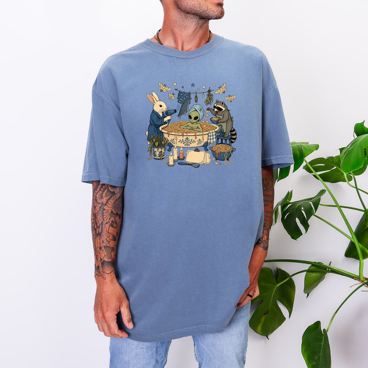 A man wearing an oversized blue t-shirt featuring an illustration of an alien bathing in spaghetti, attended by a rabbit and raccoon.