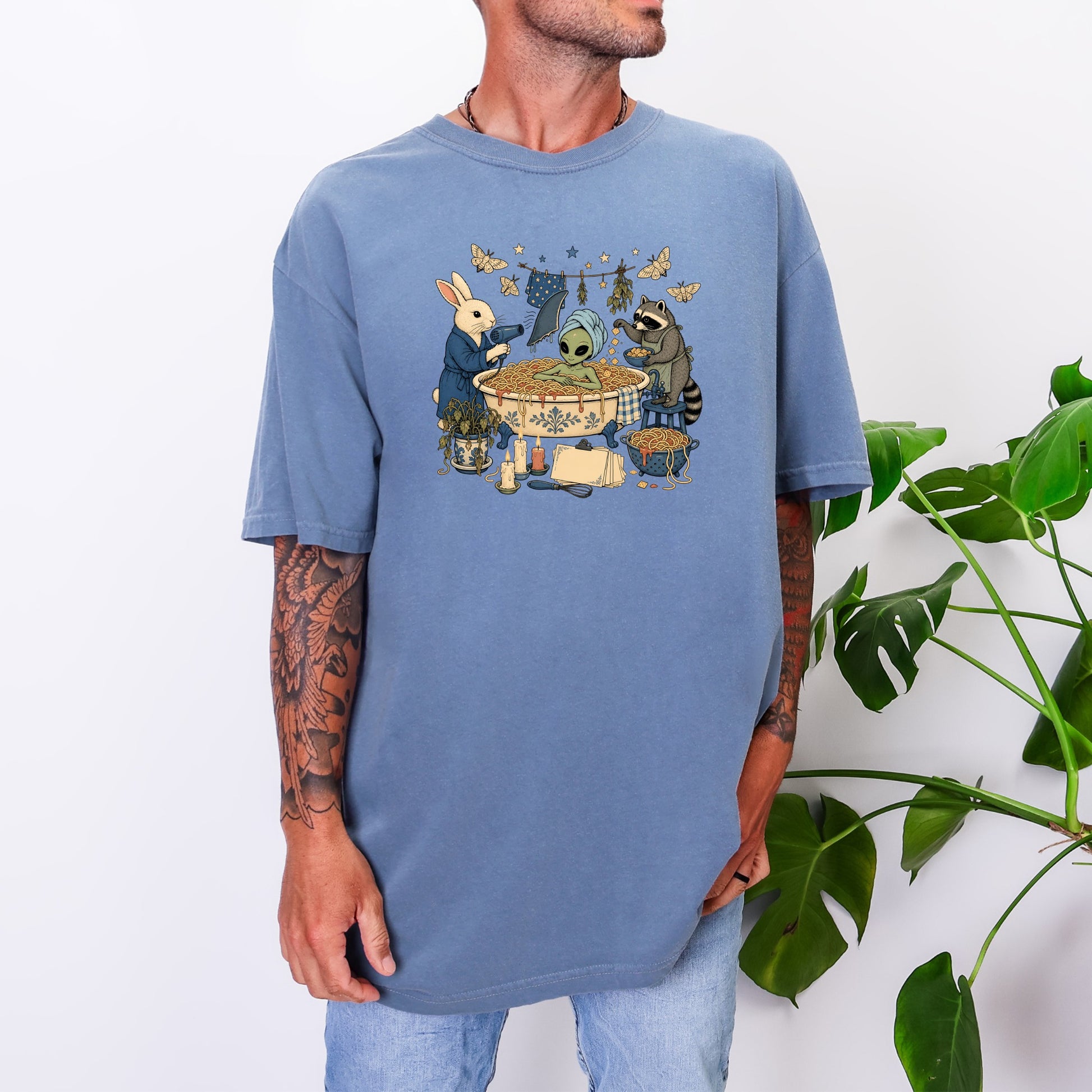 A man wearing an oversized blue t-shirt featuring an illustration of an alien bathing in spaghetti, attended by a rabbit and raccoon.