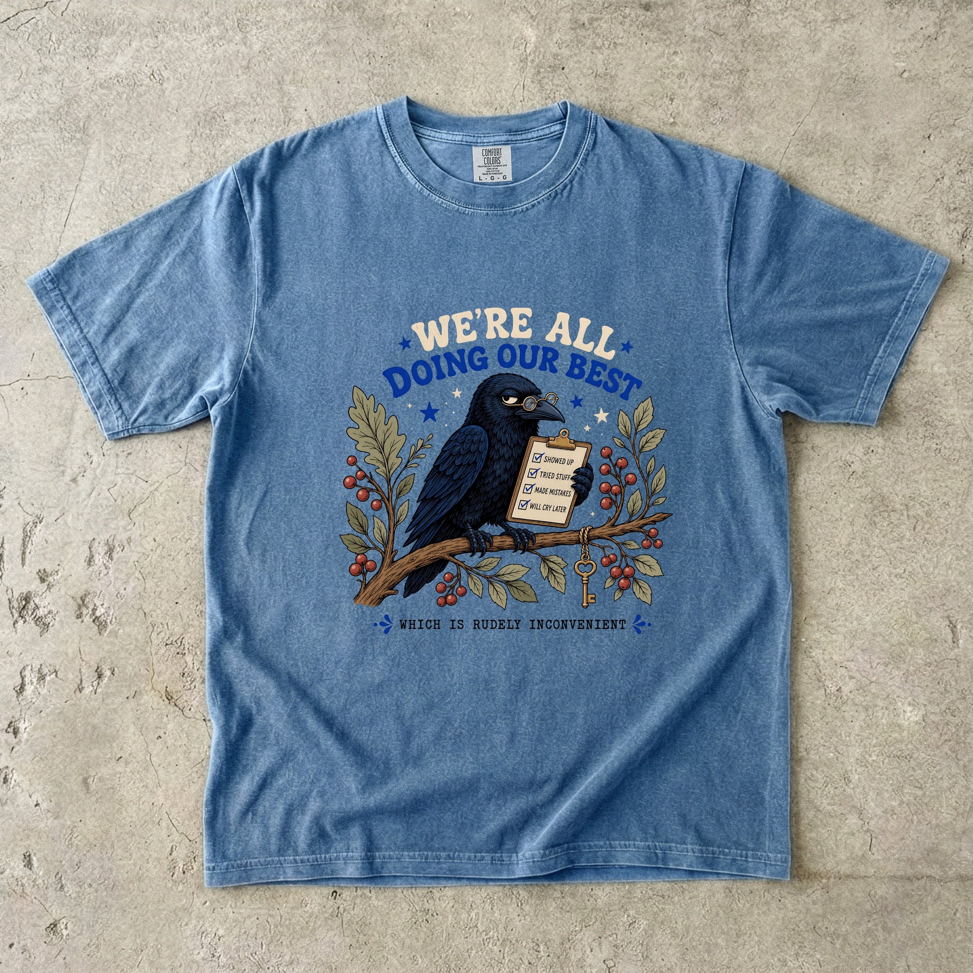 Blue t-shirt with crow wearing glasses holding a checklist, text says "We're All Doing Our Best Which Is Rudely Inconvenient".
