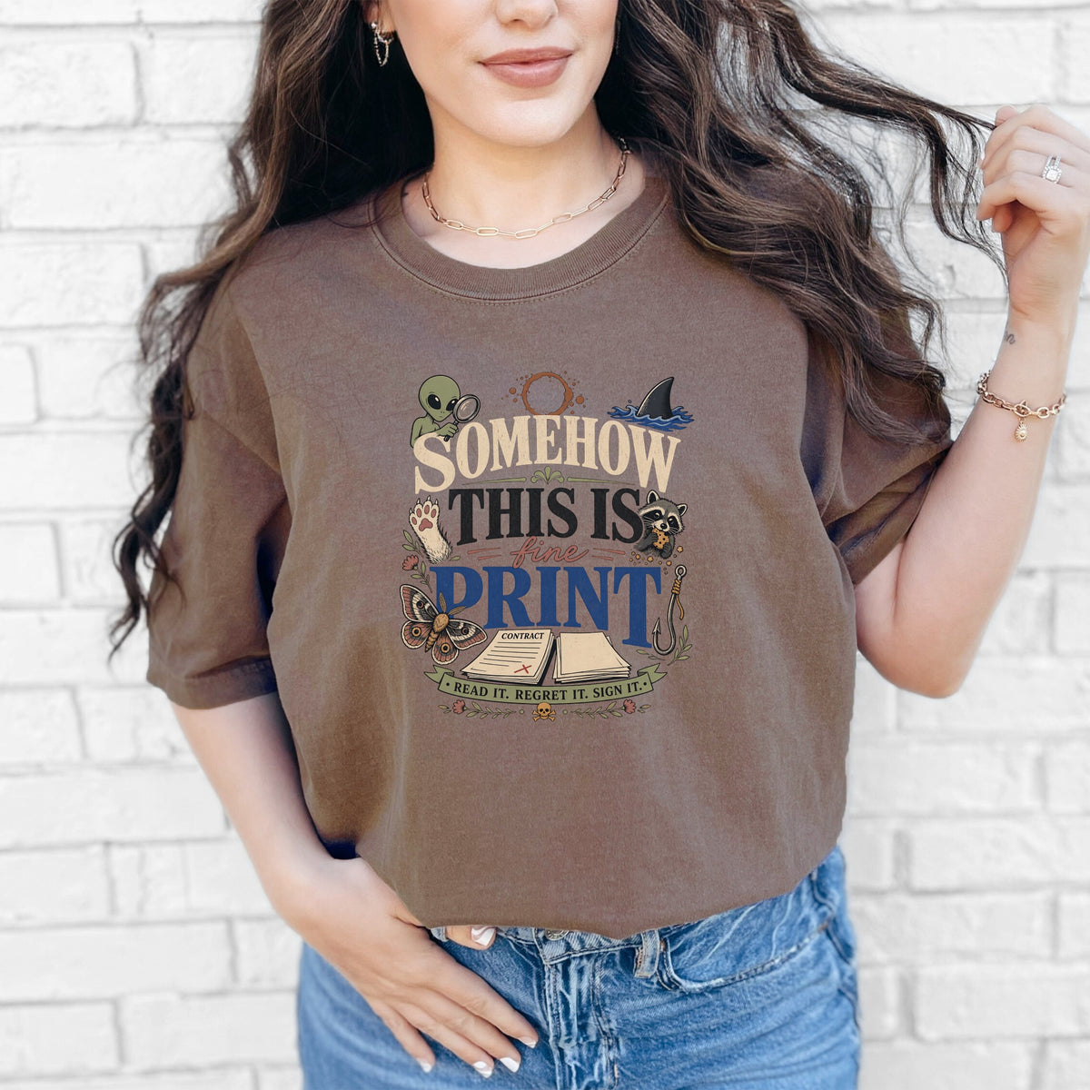 A woman wearing a brown t-shirt featuring the text "SOMEHOW THIS IS FINE PRINT" and various illustrations.