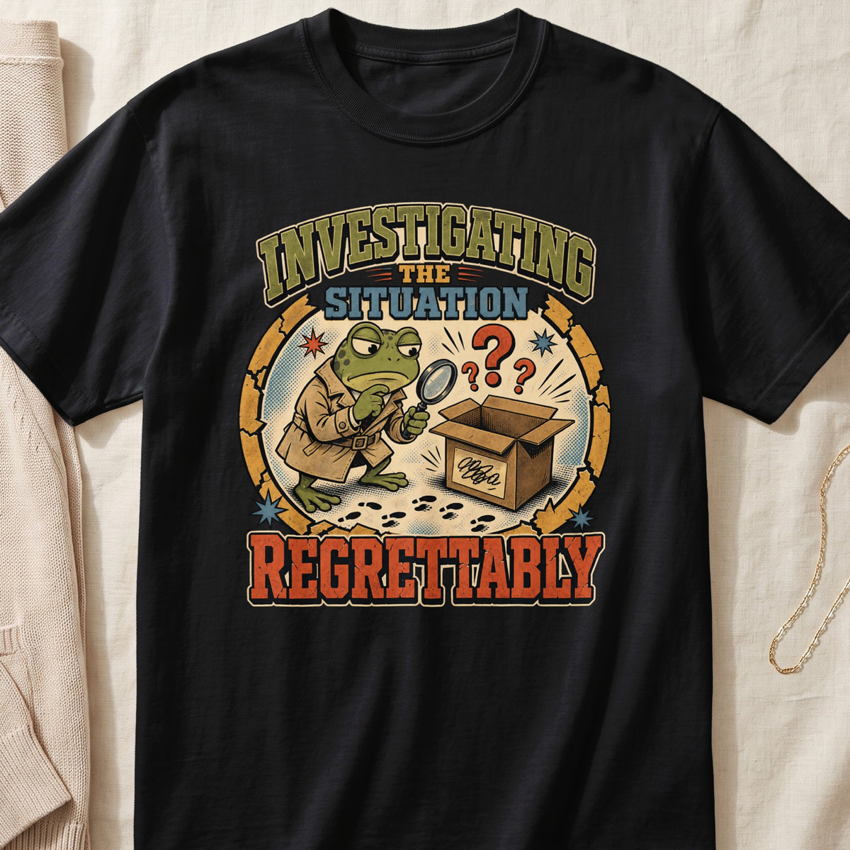 A black t-shirt features a graphic of a frog detective inspecting an open box.