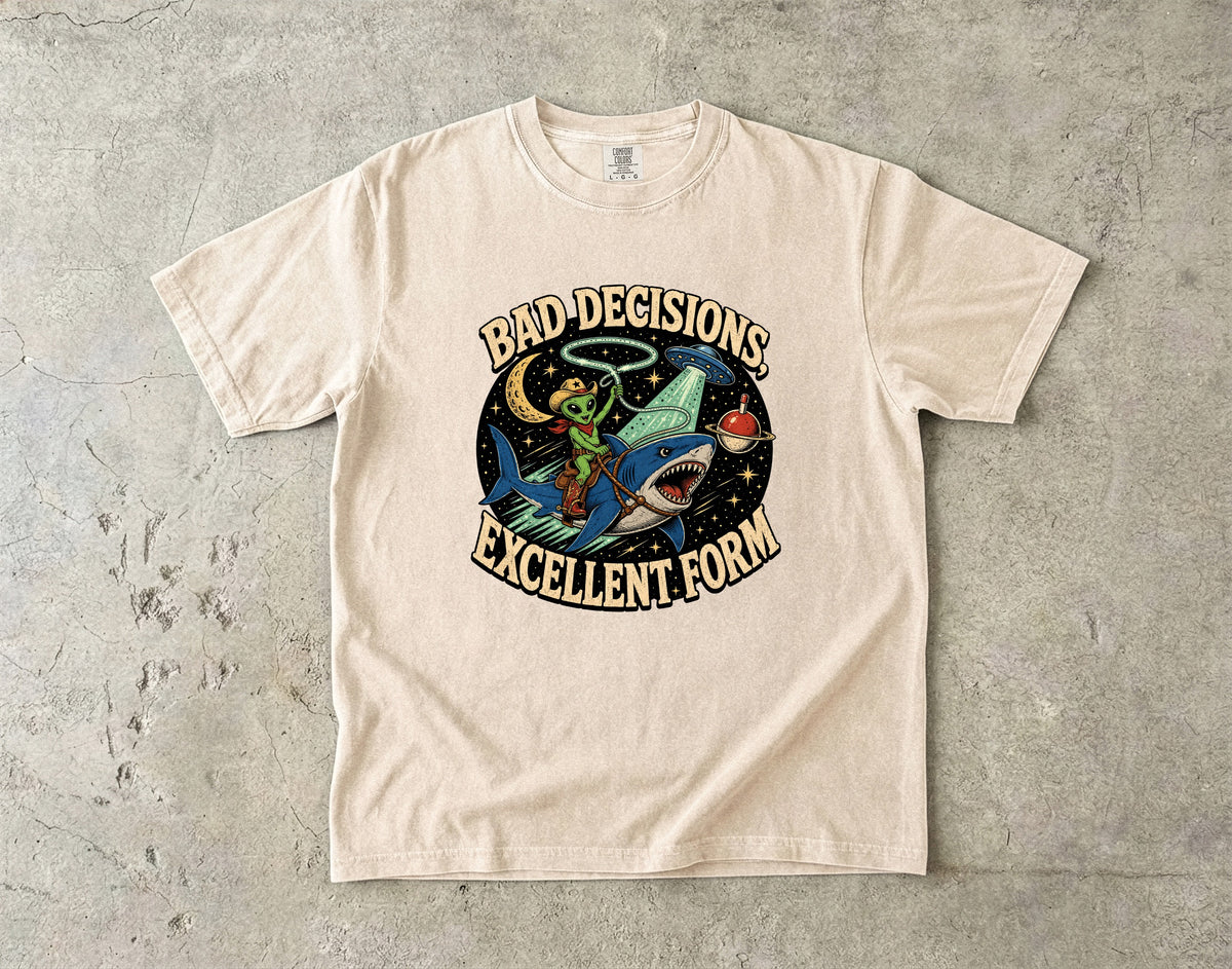 Light tan t-shirt with graphic: alien cowboy riding shark, text reads "BAD DECISIONS, EXCELLENT FORM".