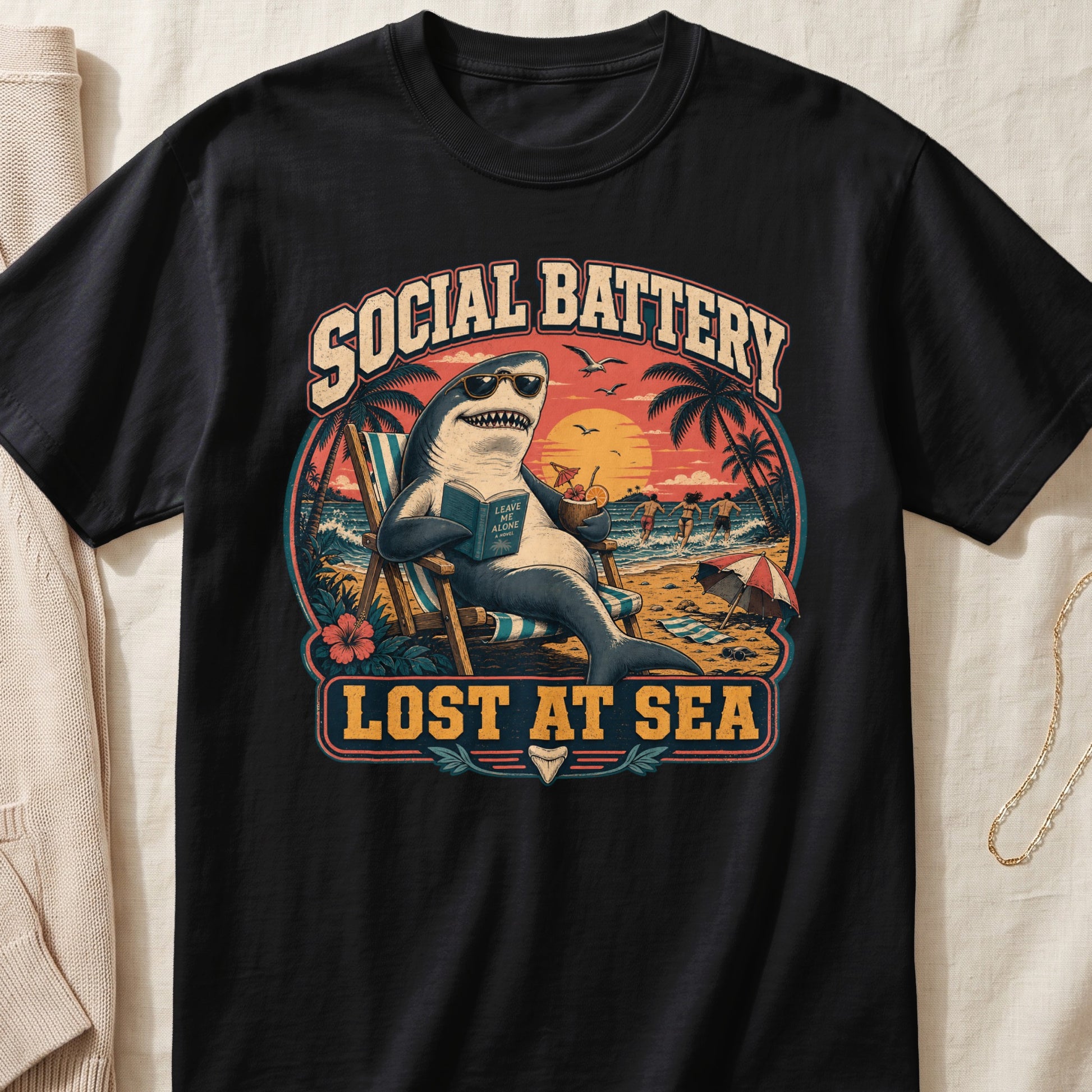 Shark wearing sunglasses reading a book on a beach with text "SOCIAL BATTERY LOST AT SEA".