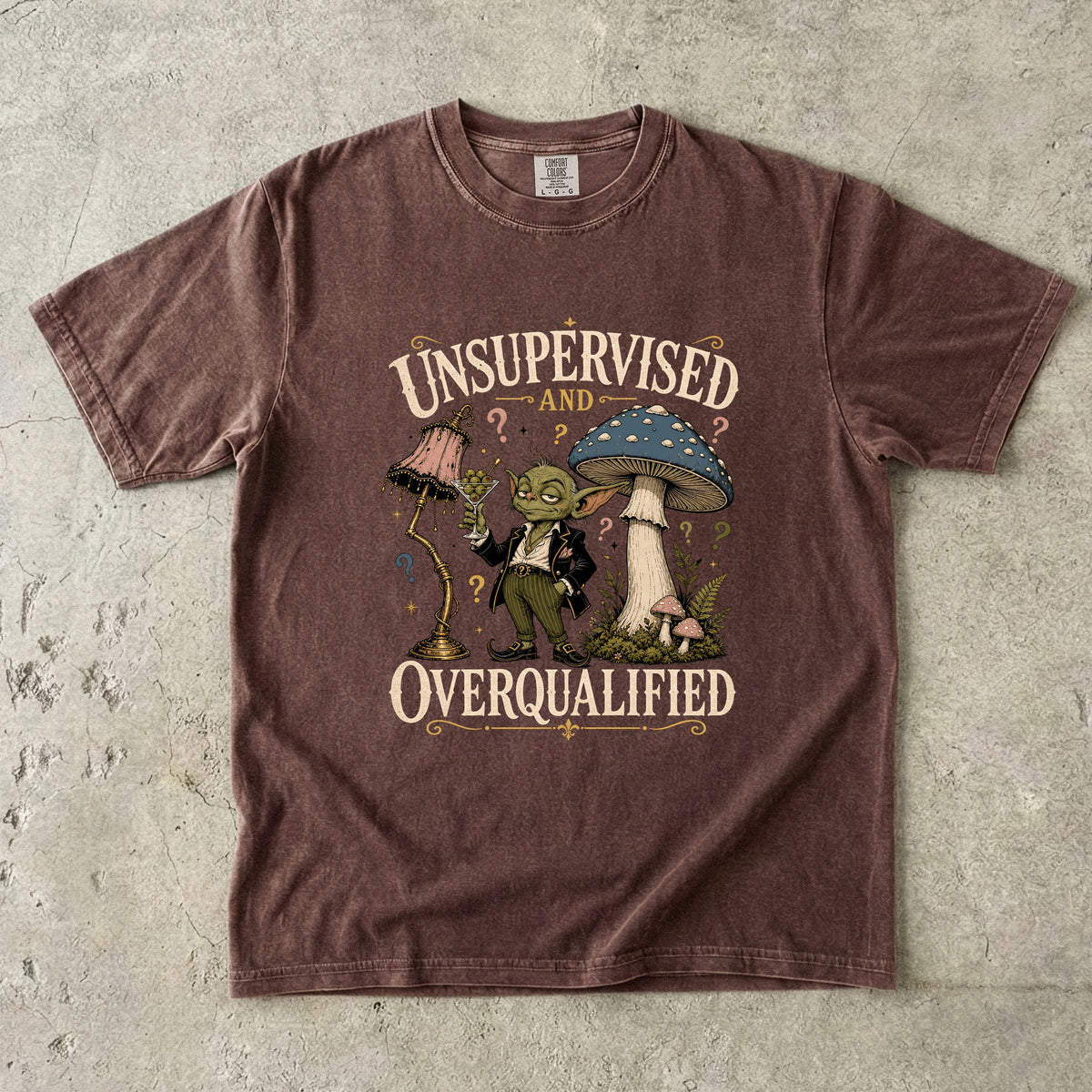 Brown t-shirt with illustration of a character standing between a lamp and a mushroom, text reads "UNSUPERVISED AND OVERQUALIFIED".