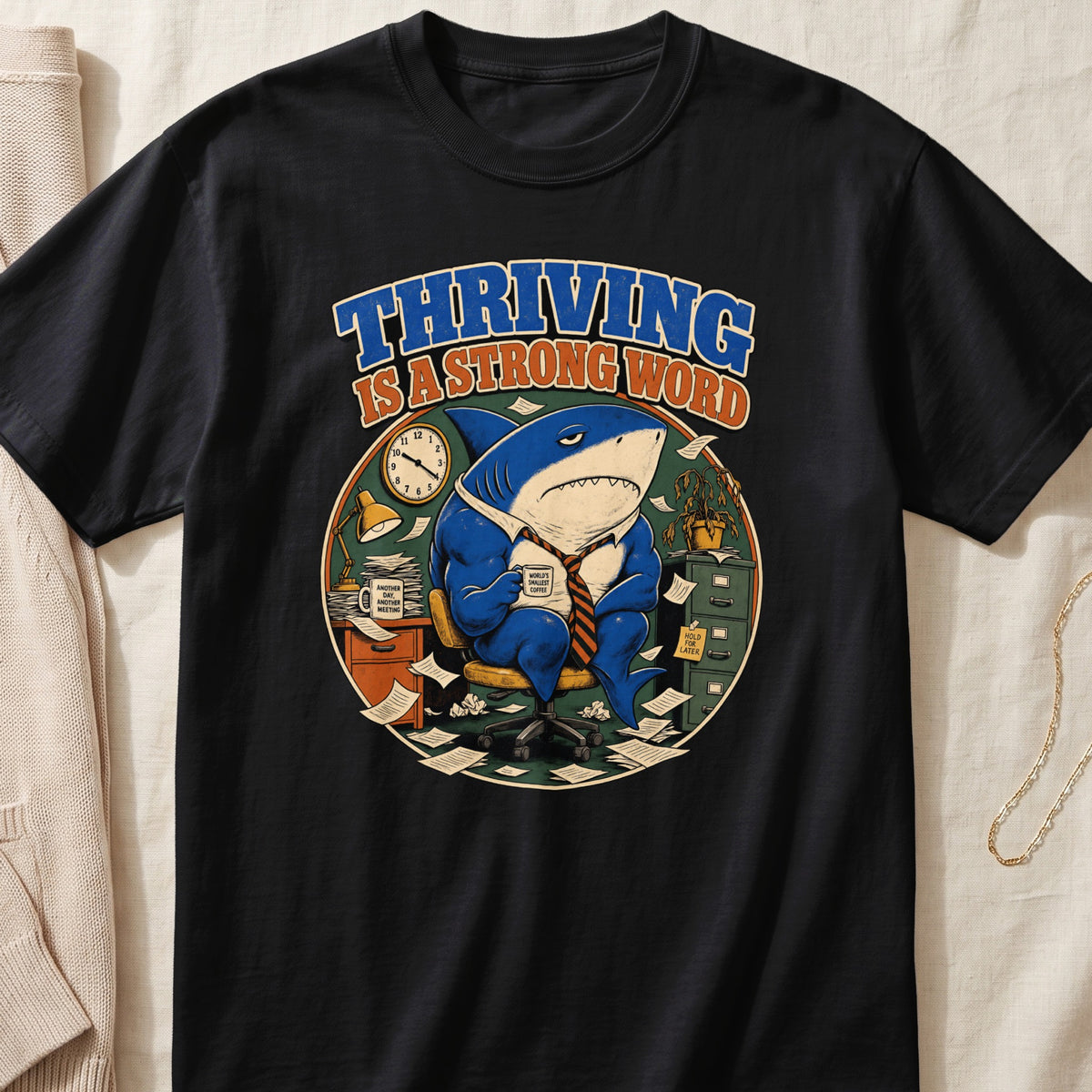 Black t-shirt with a shark in an office, drinking coffee, reading "Thriving is a strong word."
