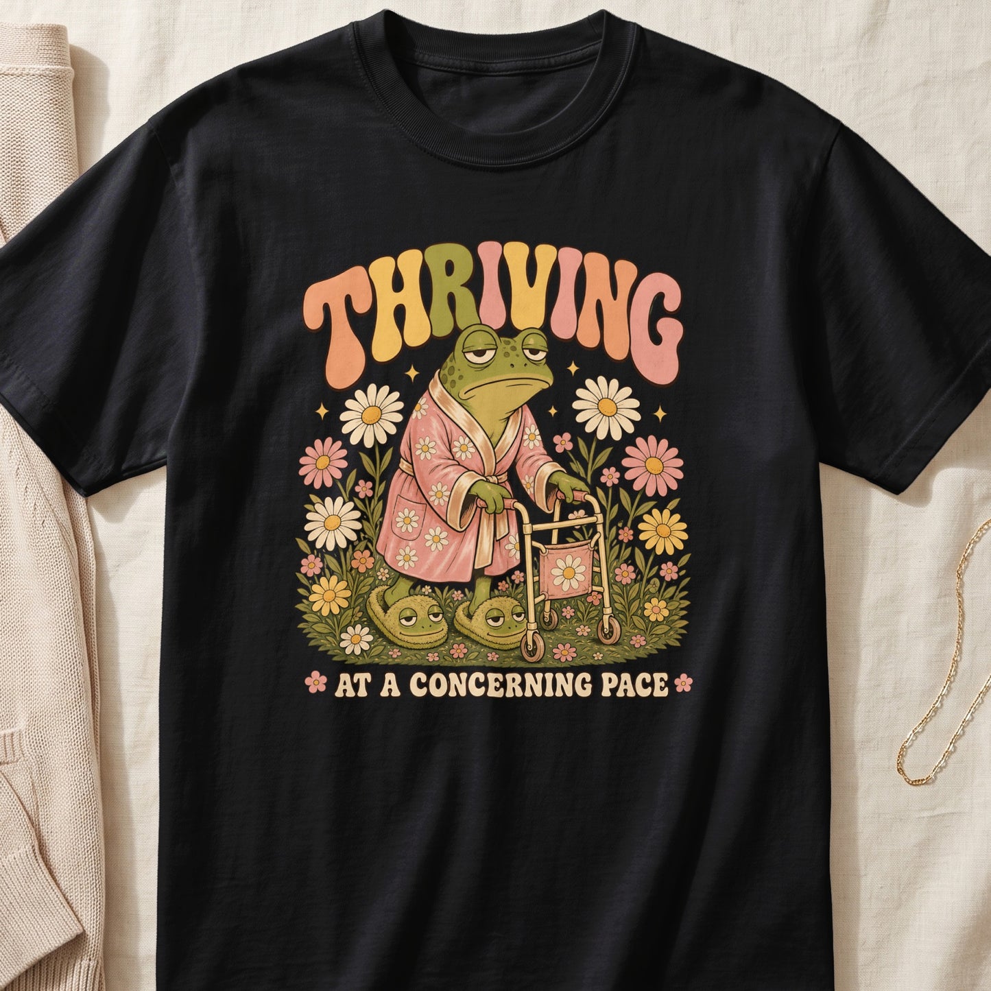 Black t-shirt with a graphic of a frog in a bathrobe and slippers using a walker, surrounded by flowers. Text reads "THRVING AT A CONCERNING PACE".