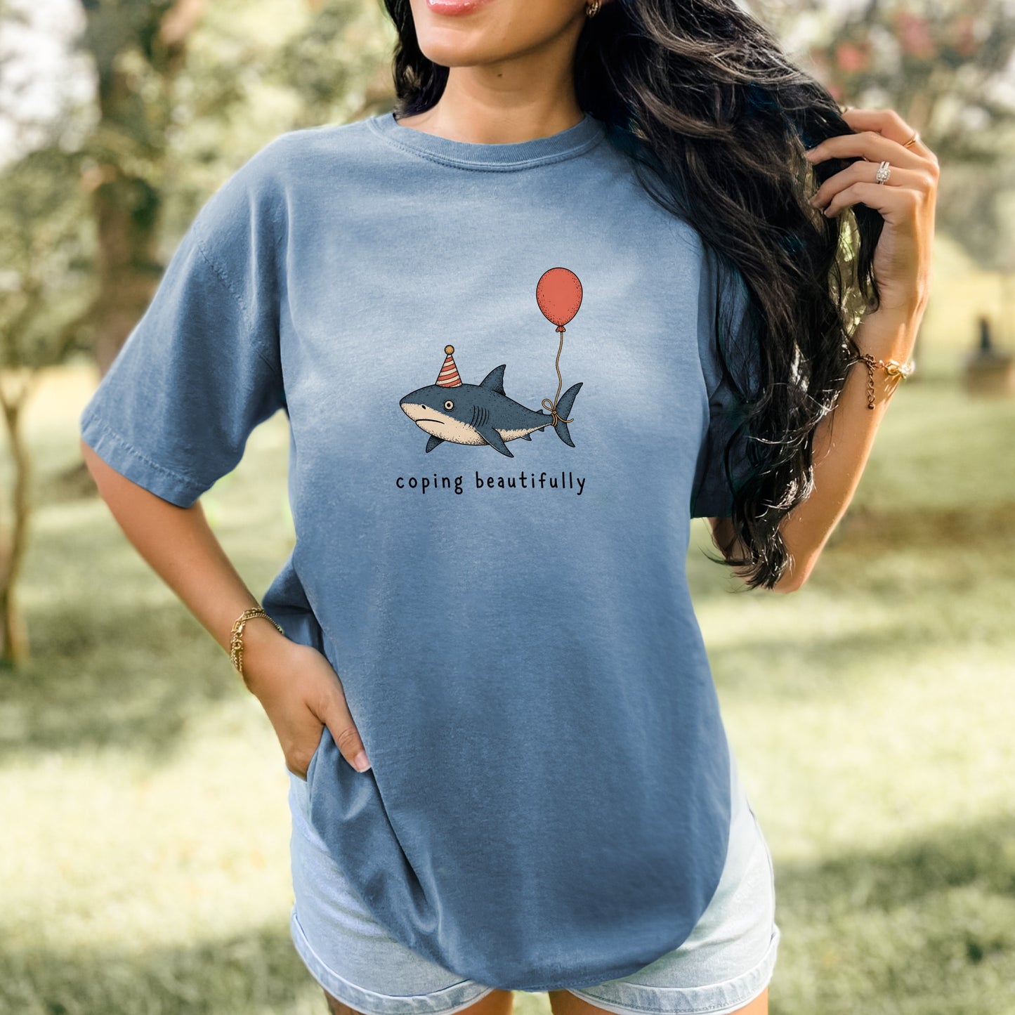 T-shirt with illustration of a shark wearing a party hat holding a balloon, with text "coping beautifully".