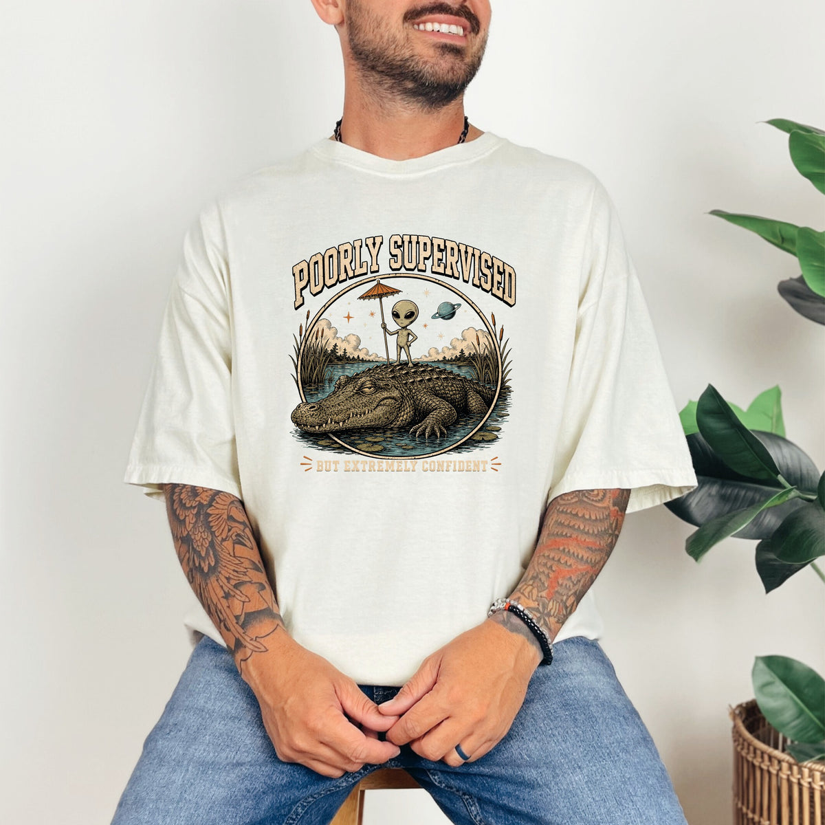 T-shirt graphic with text "POORLY SUPERVISED BUT EXTREMELY CONFIDENT" featuring an alien on an alligator.