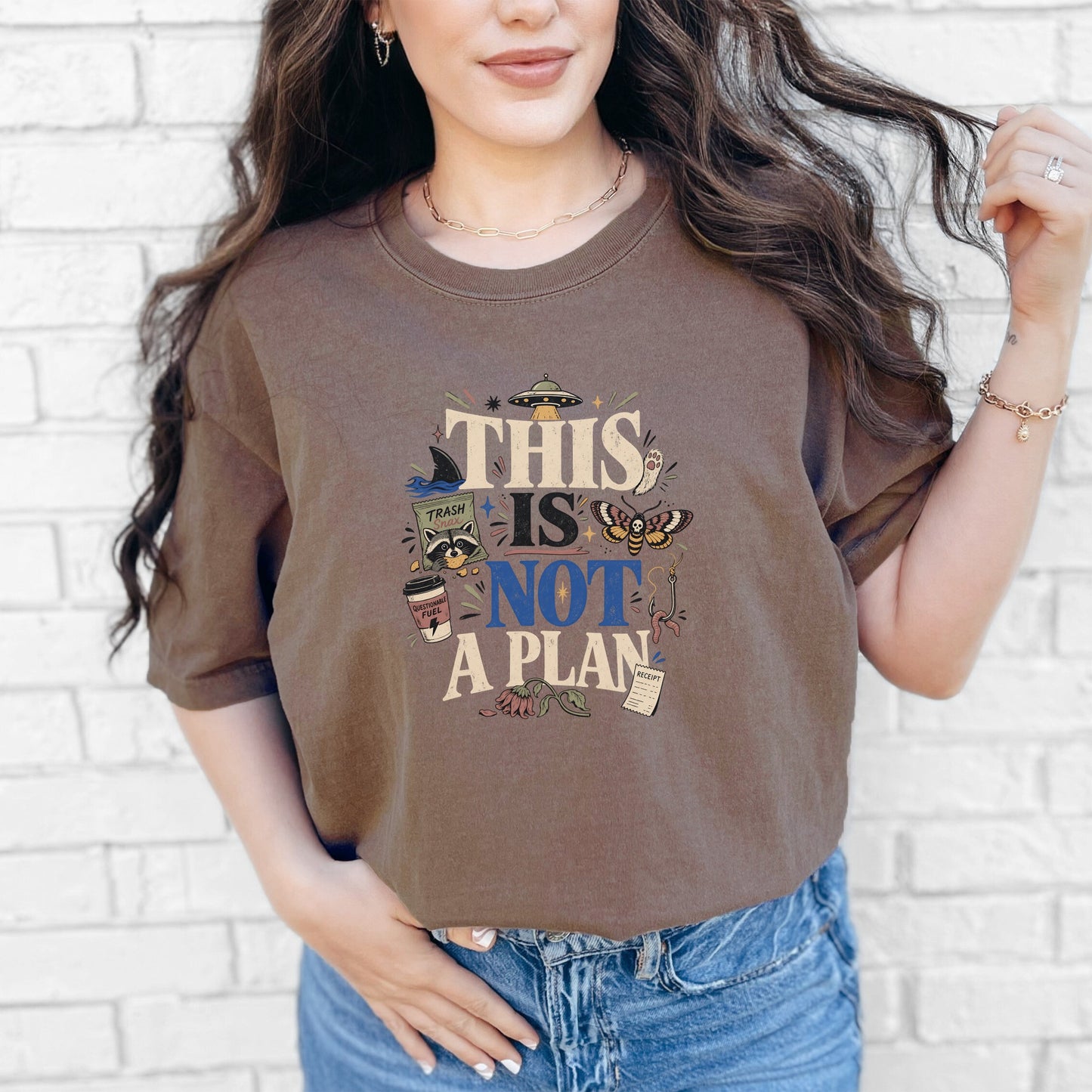 A person wearing a brown crewneck t-shirt featuring the text "THIS IS NOT A PLAN" and quirky illustrations.