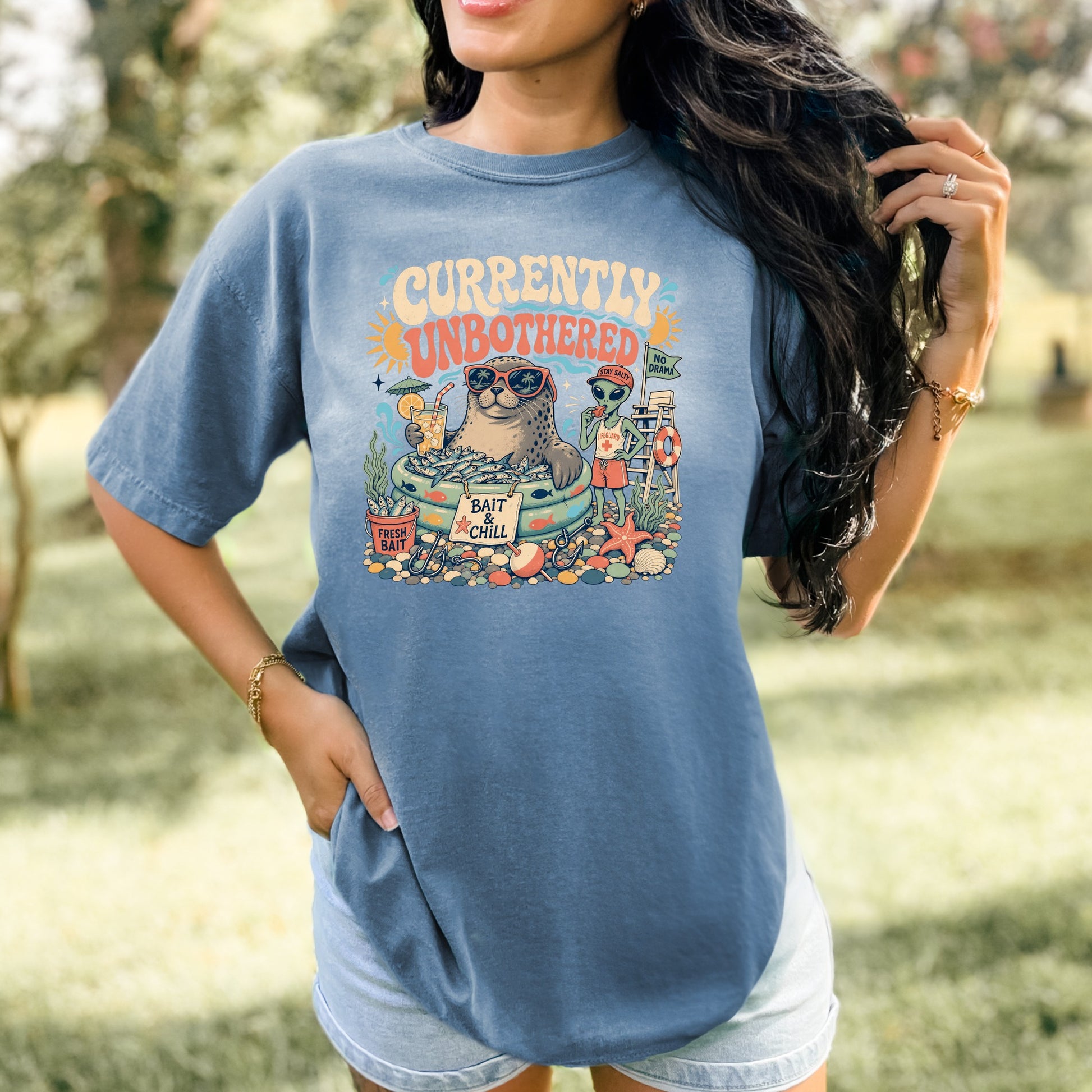 Graphic tee with seal in kiddie pool, alien lifeguard, and text "Currently Unbothered".