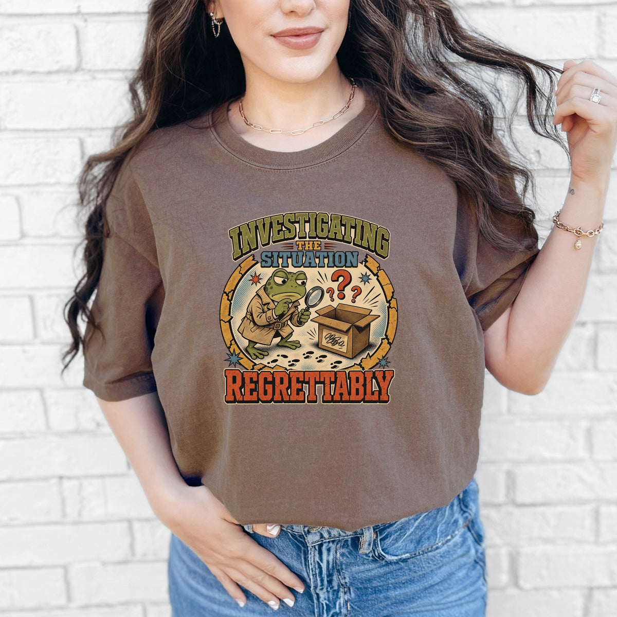 A woman wearing a brown t-shirt featuring a cartoon frog in a trench coat examining a box with a magnifying glass.