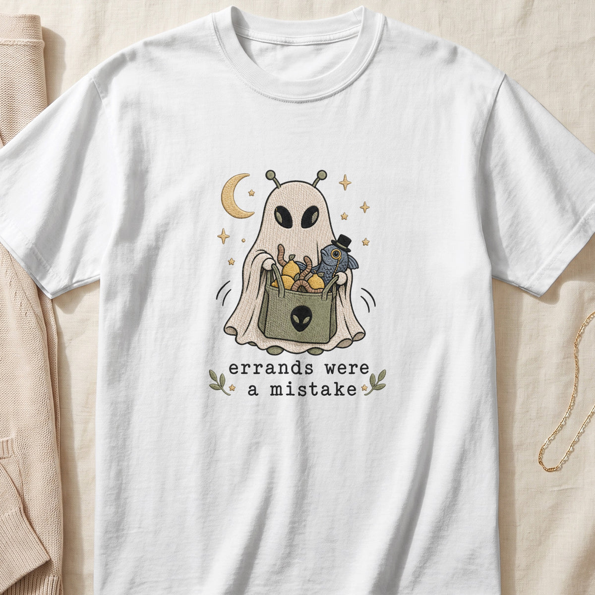 White t-shirt featuring an embroidered illustration of an alien in a bedsheet ghost costume holding a grocery bag, with the text "errands were a mistake".