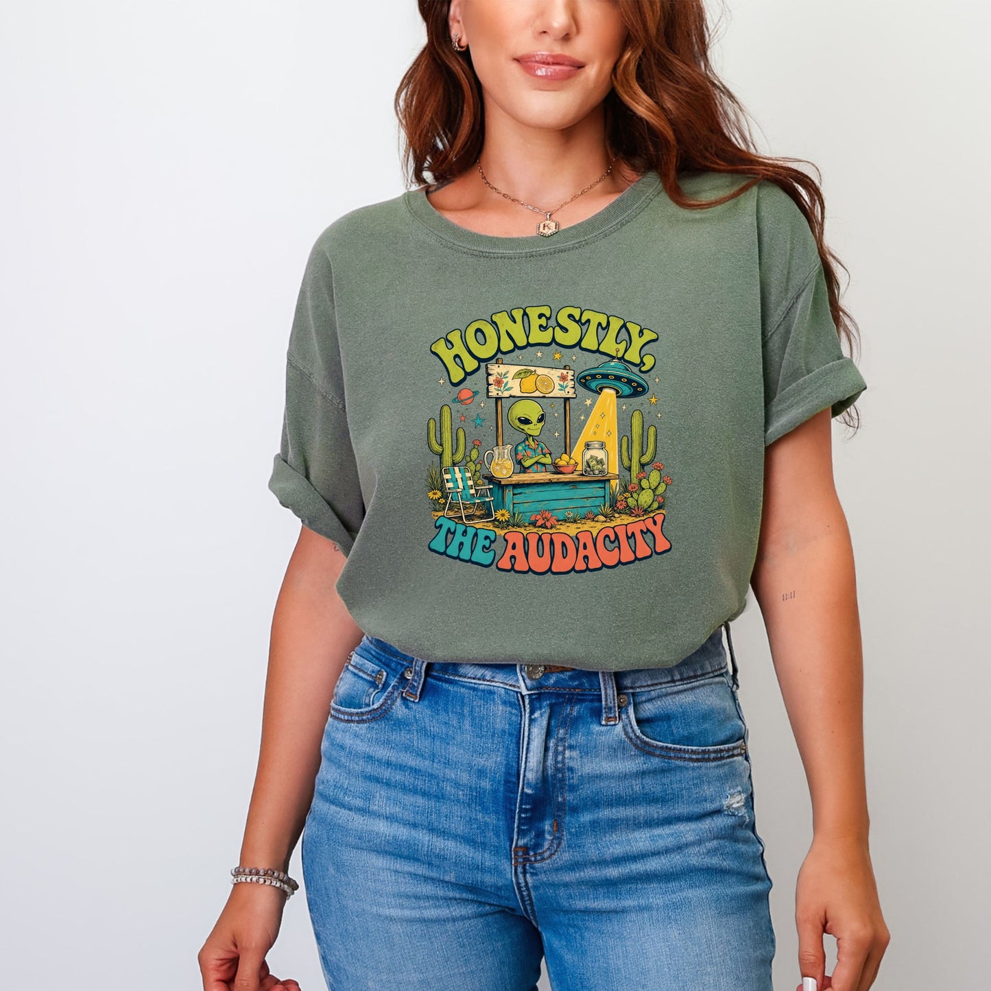 Green t-shirt with graphic of alien at lemonade stand and text "HONESTLY THE AUDACITY."