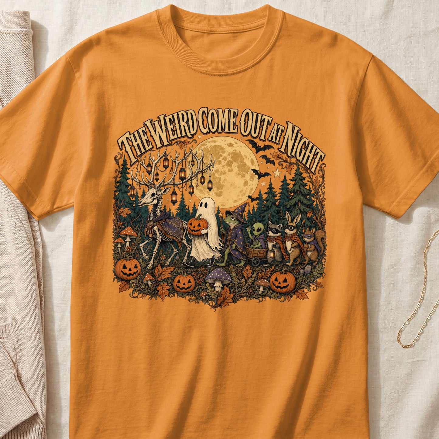 Orange t-shirt with Halloween graphic: "The Weird Come Out At Night" text, skeleton deer, ghost, frog wizard, alien, raccoon, rabbit, and pumpkins.