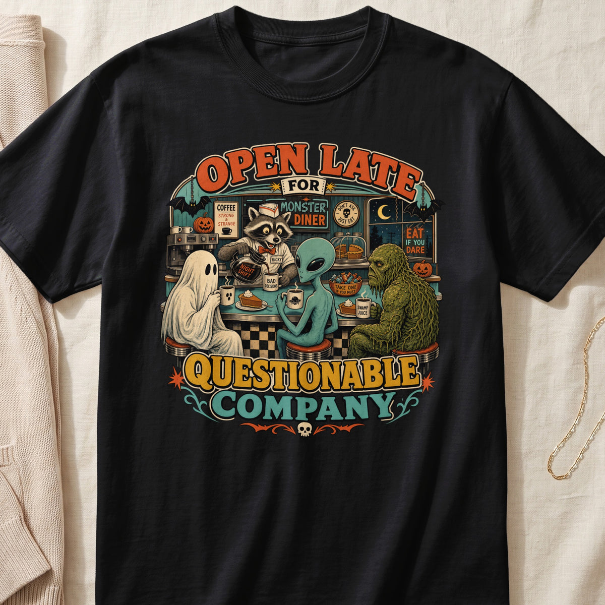 Black t-shirt featuring a retro diner scene with a ghost, alien, raccoon, and swamp monster.