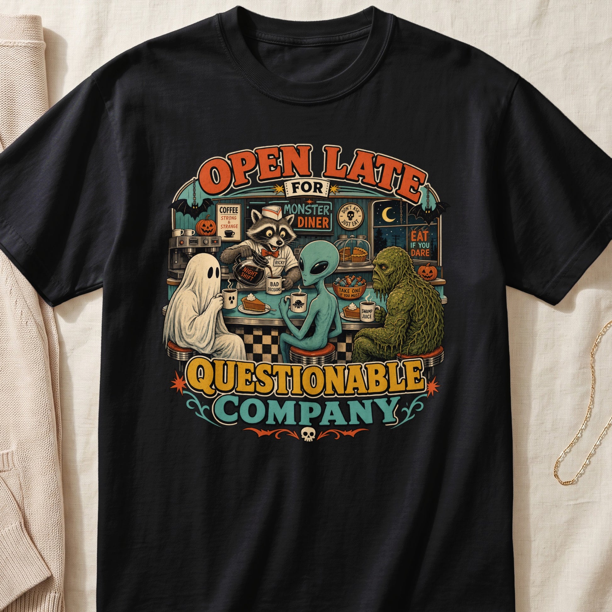 Black t-shirt featuring a retro diner scene with a ghost, alien, raccoon, and swamp monster.