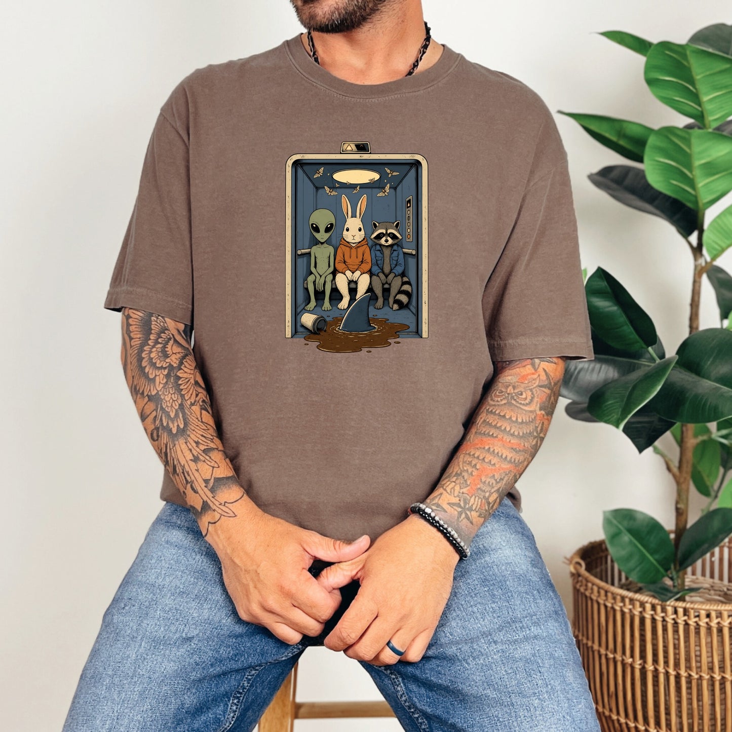 A tattooed man wearing a brown t-shirt featuring an alien, rabbit, and raccoon inside an elevator.