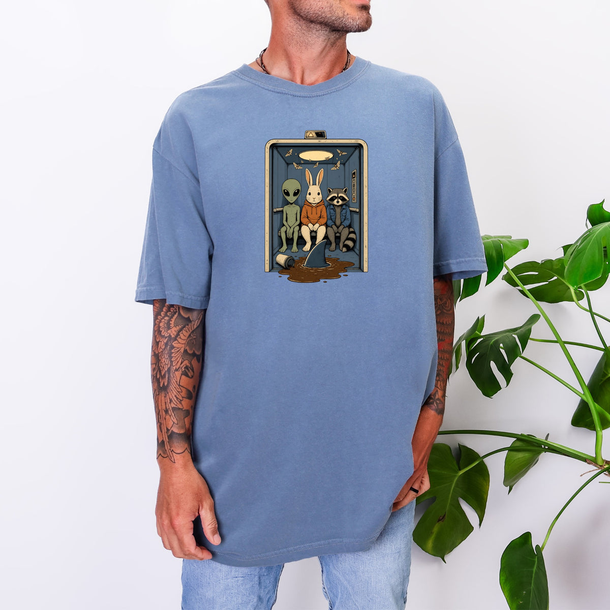 A tattooed man wearing a blue oversized t-shirt with an alien, rabbit, and raccoon graphic.