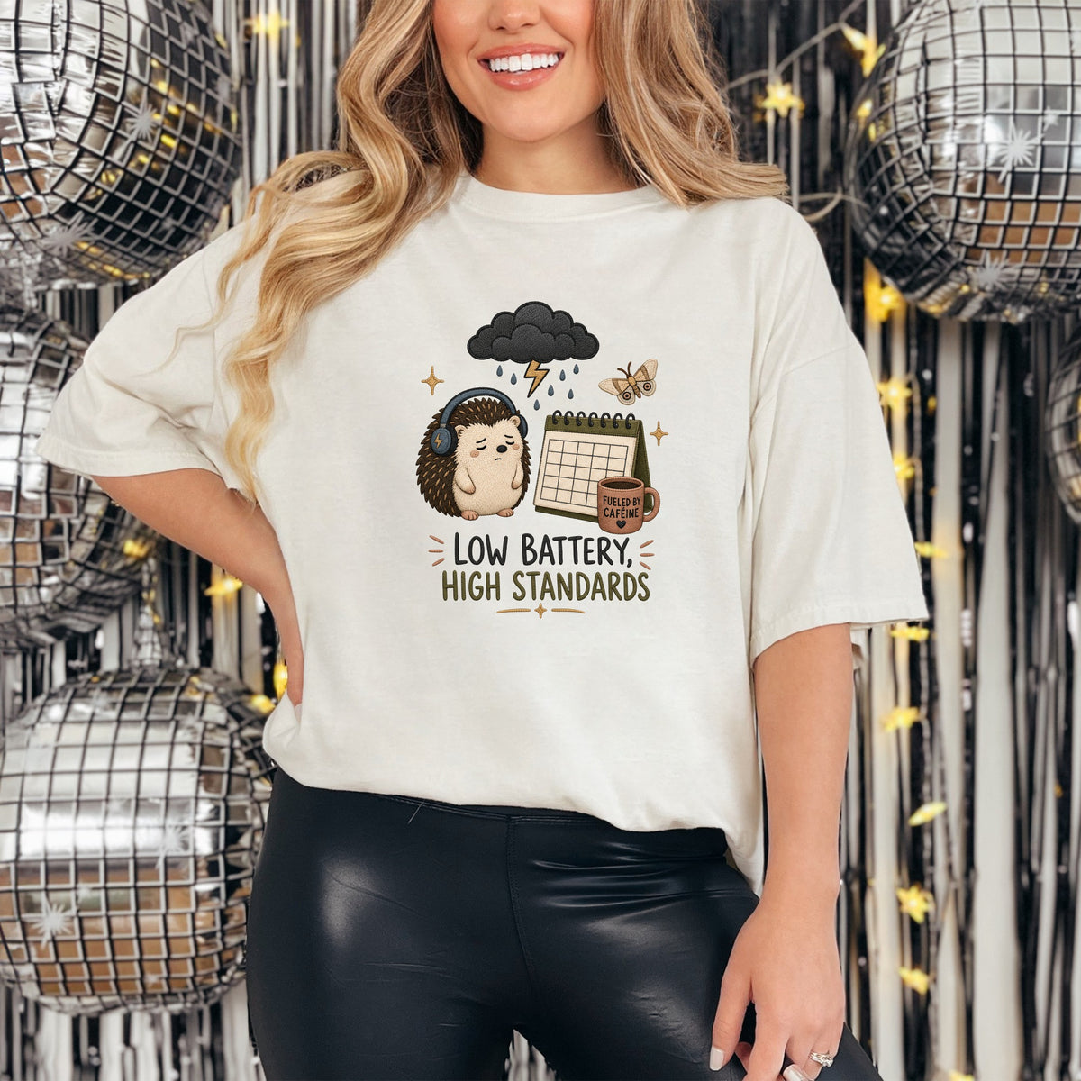 A woman wearing a white t-shirt featuring an embroidered sad hedgehog, rainy cloud, and calendar graphic with the text "LOW BATTERY, HIGH STANDARDS".