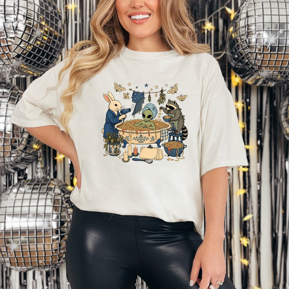 A woman wearing a white t-shirt featuring a graphic of an alien bathing in spaghetti, attended by a rabbit and raccoon.