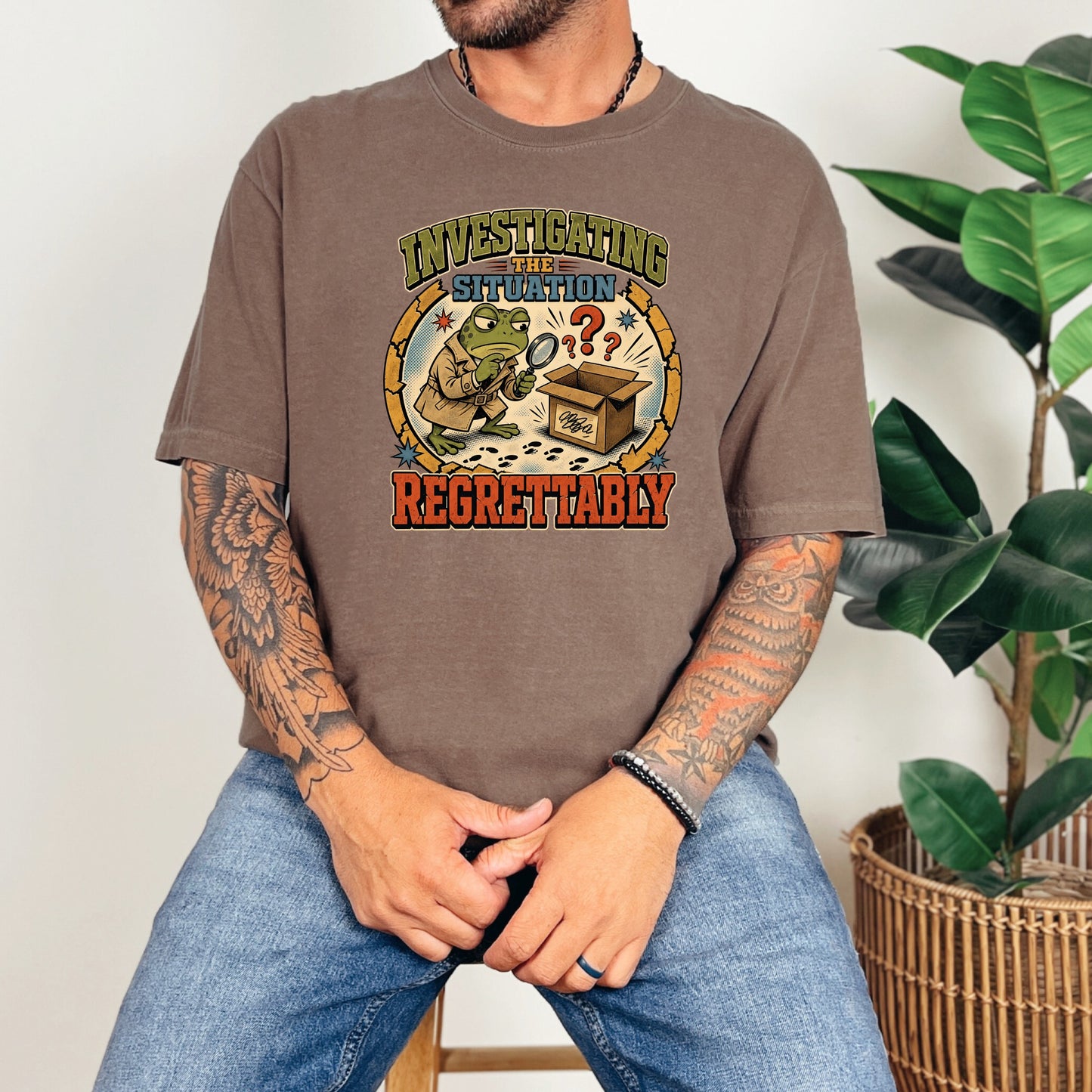 A person wearing a brown t-shirt featuring a cartoon frog holding a magnifying glass and text reading Investigating the Situation Regrettably.