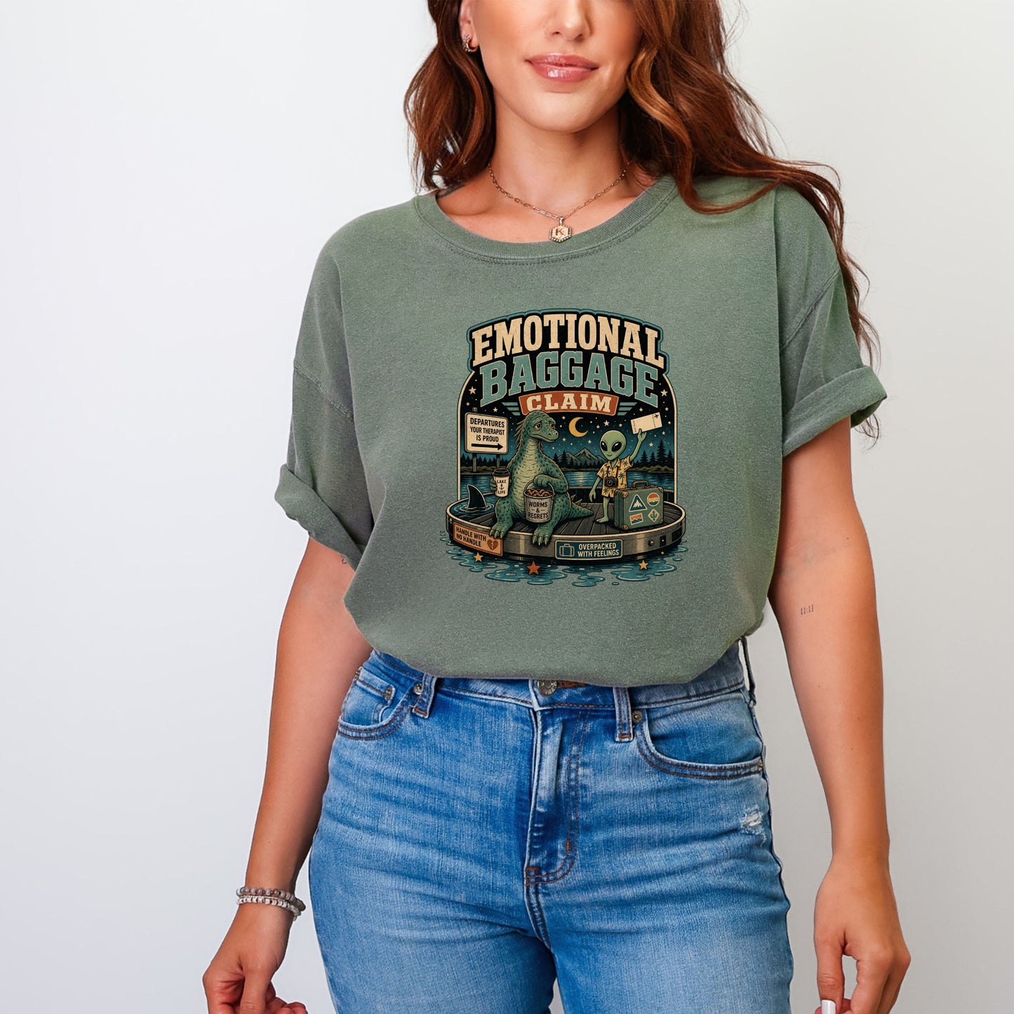 Graphic on a green t-shirt with "Emotional Baggage Claim," a dinosaur, and an alien.