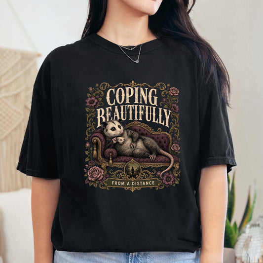 Black t-shirt with an opossum on a couch, text "COPING BEAUTIFULLY FROM A DISTANCE".