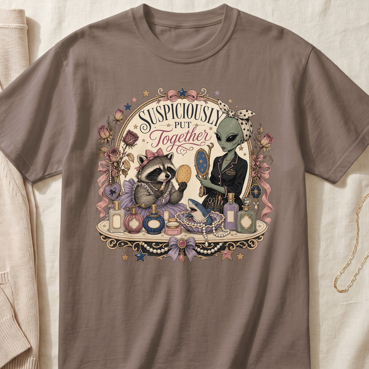 A grey t-shirt featuring a retro graphic of an alien and a raccoon applying makeup.