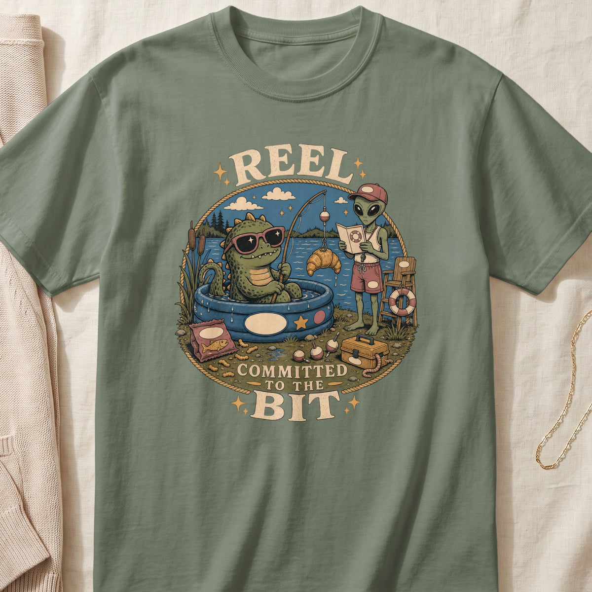 Green t-shirt with "REEL COMMITTED TO THE BIT" text and cartoon of alien and dinosaur fishing.