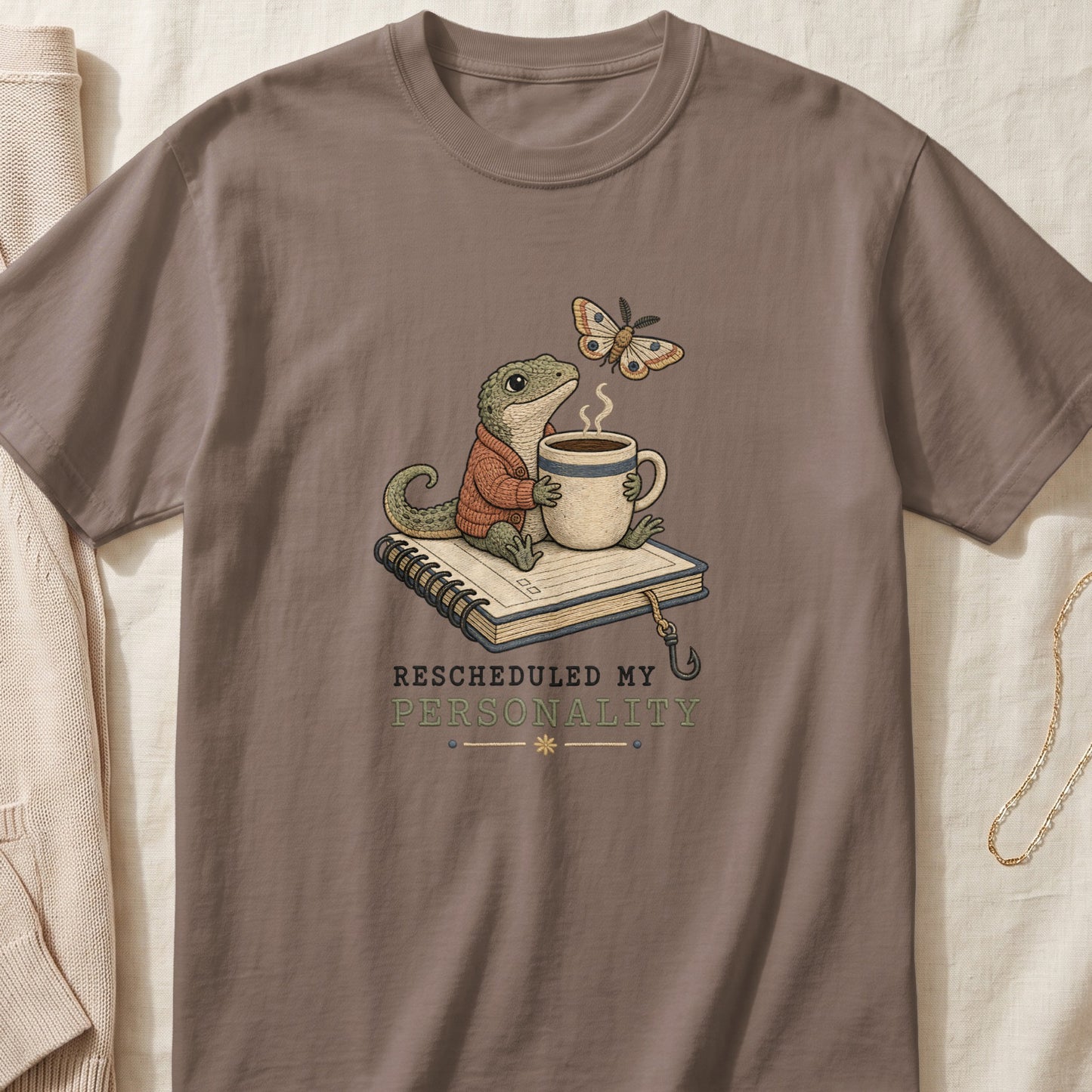 A brown t-shirt featuring an illustration of a lizard holding a coffee mug while sitting on a notebook, accompanied by a moth and text reading "Rescheduled My Personality."