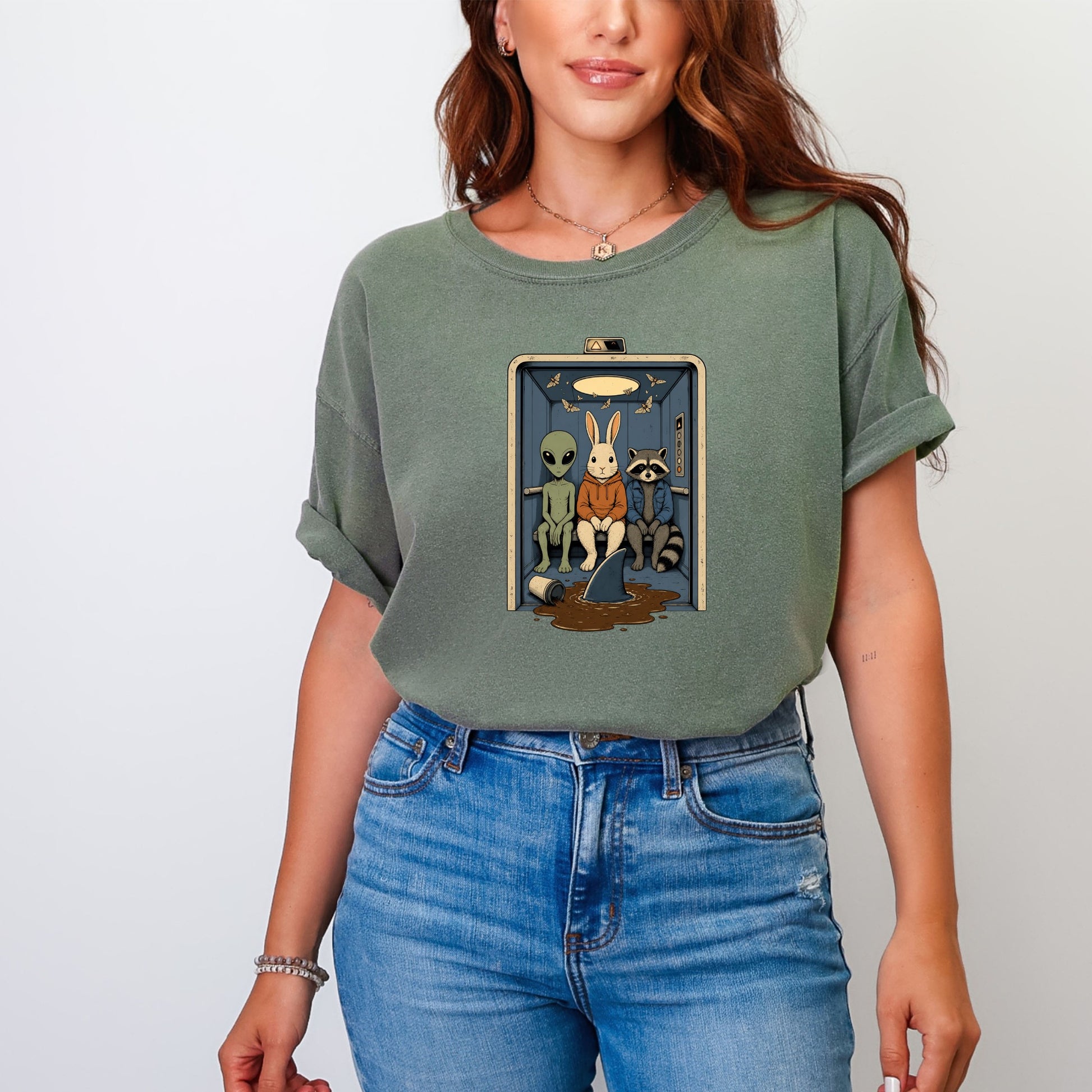 A woman wearing a faded green t-shirt featuring an illustration of an alien, a rabbit, and a raccoon riding in an elevator.