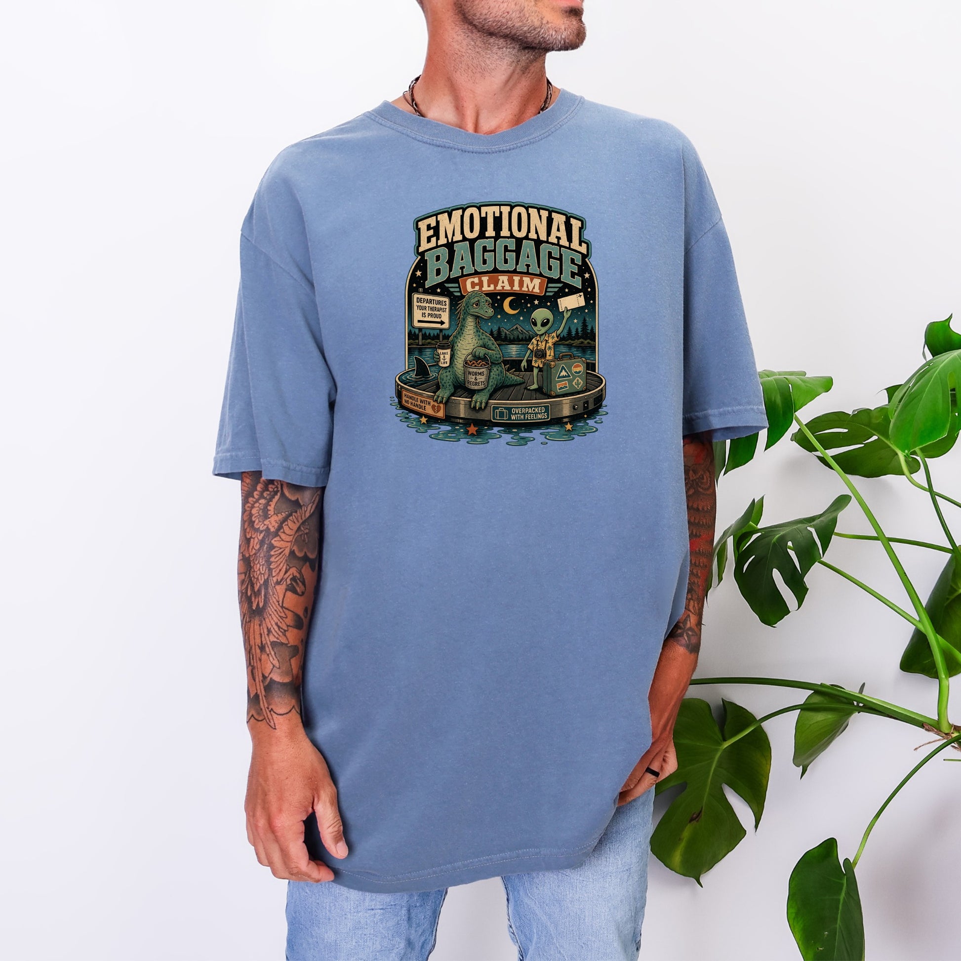 T-shirt with "Emotional Baggage Claim" text, dinosaur, alien, and suitcase graphic.