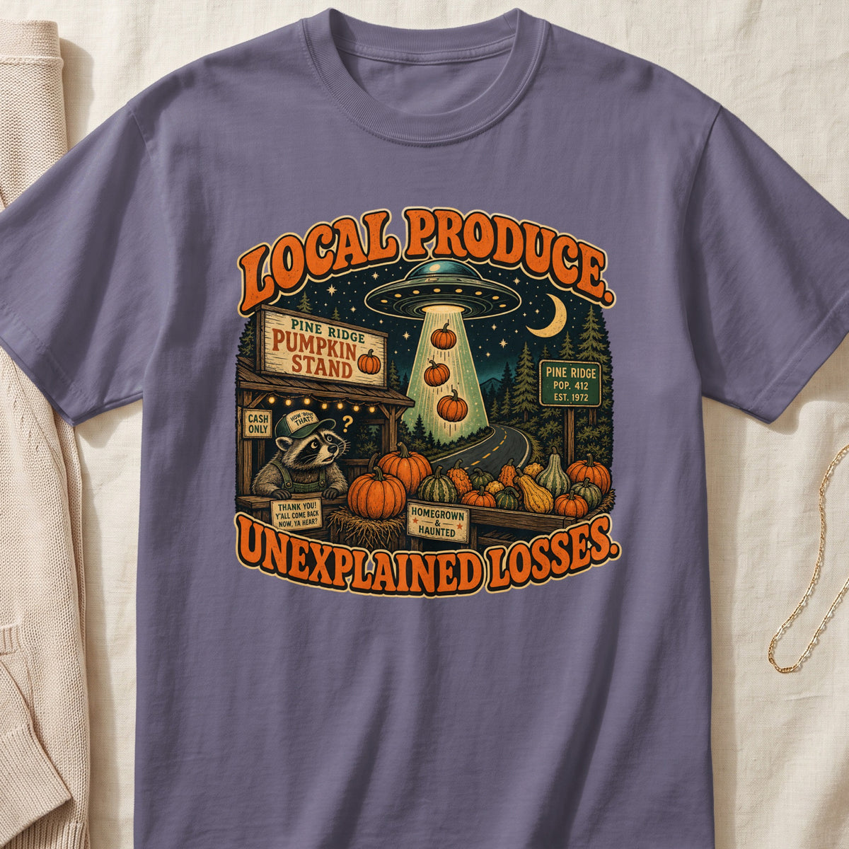 Graphic tee with "Local Produce Unexplained Losses" text, a raccoon, pumpkins, and a UFO.