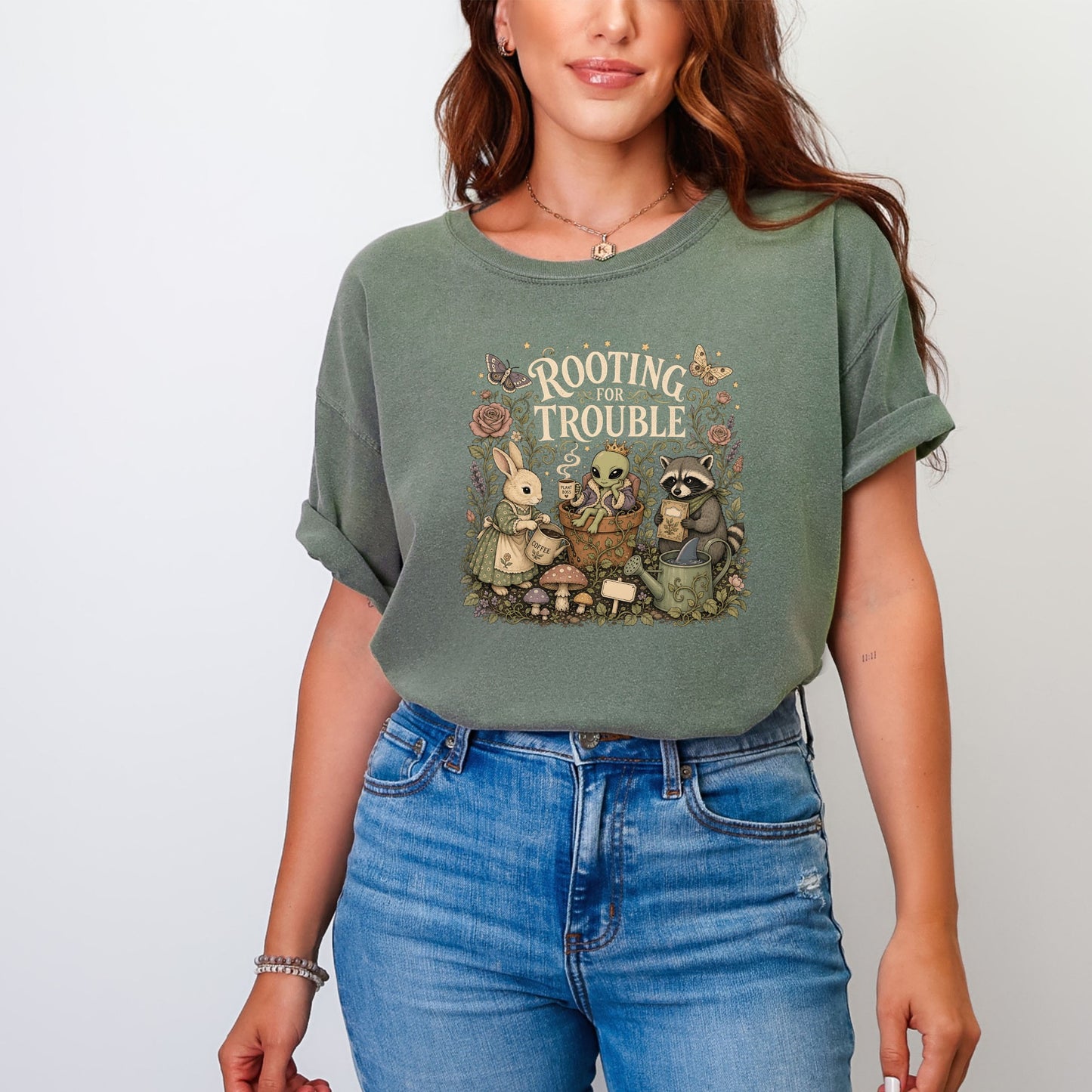 Green t-shirt graphic with text "Rooting for Trouble", a bunny, alien, and raccoon in a garden scene.
