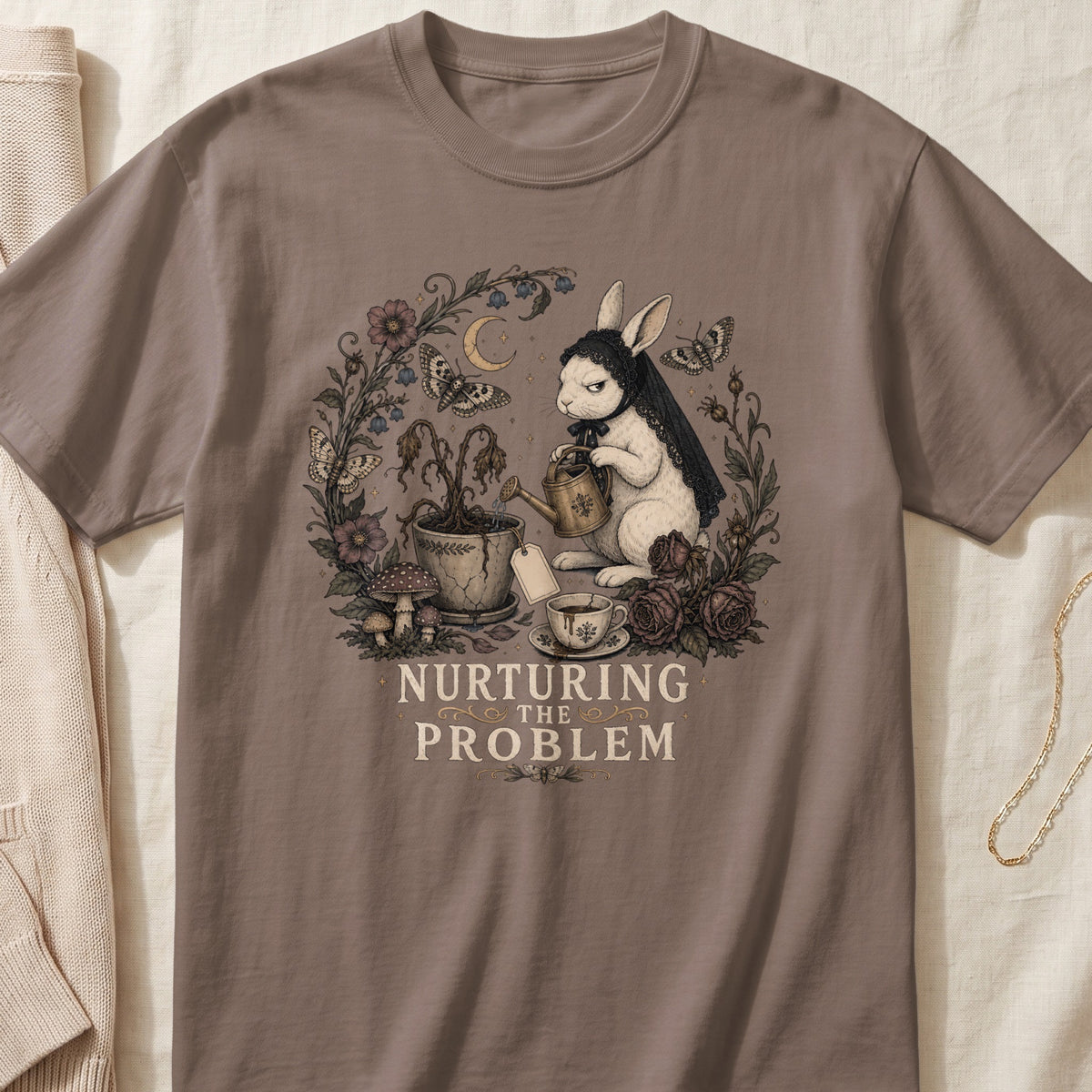 A brown t-shirt featuring a graphic of a rabbit watering a dead plant, with text reading NURTURING THE PROBLEM.
