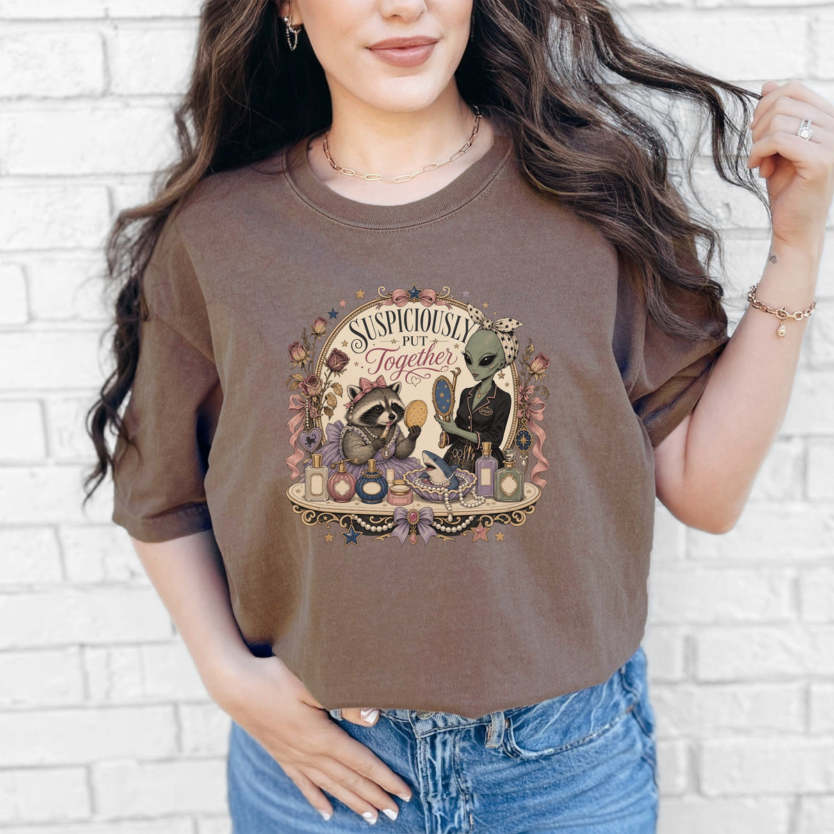 A woman wearing a brown t-shirt featuring a vintage-style graphic that reads Suspiciously Put Together with a raccoon and an alien.