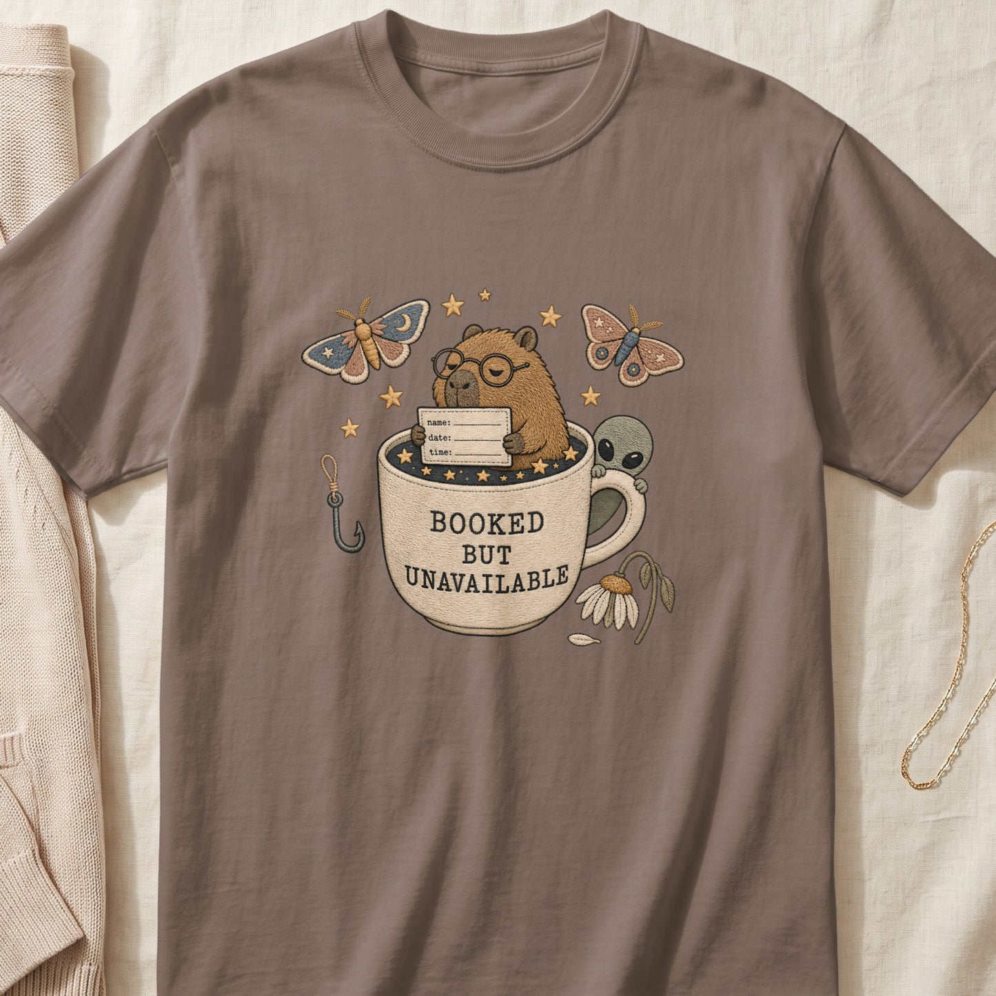 Capybara with alien in mug reading "Booked but Unavailable", with moths and fishing hook.