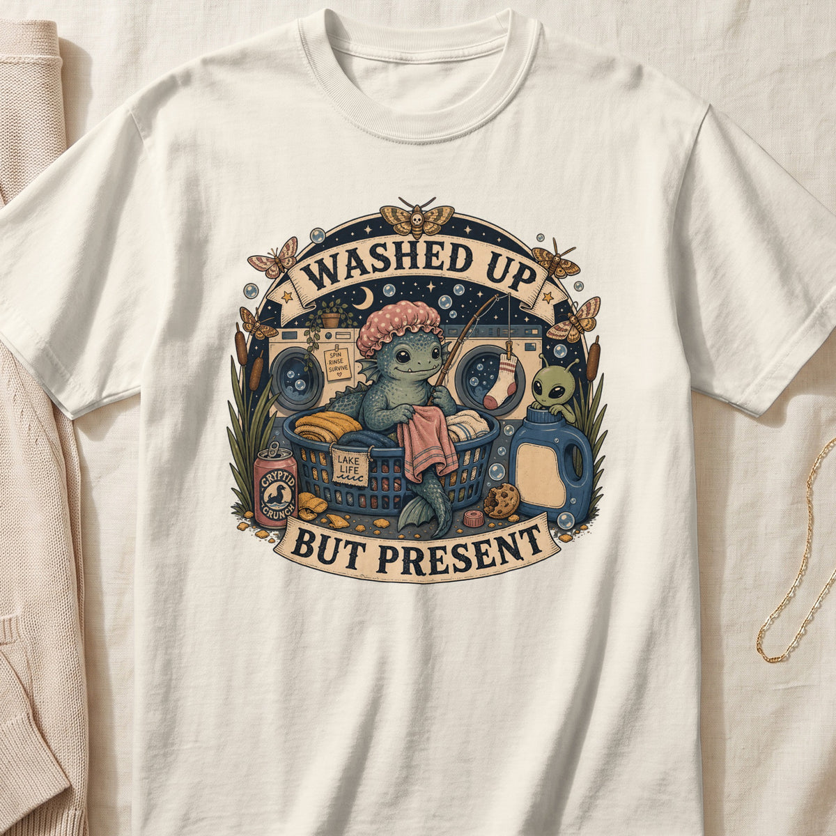 Illustration on a cream t-shirt with a cryptid character doing laundry, text says "WASHED UP BUT PRESENT".